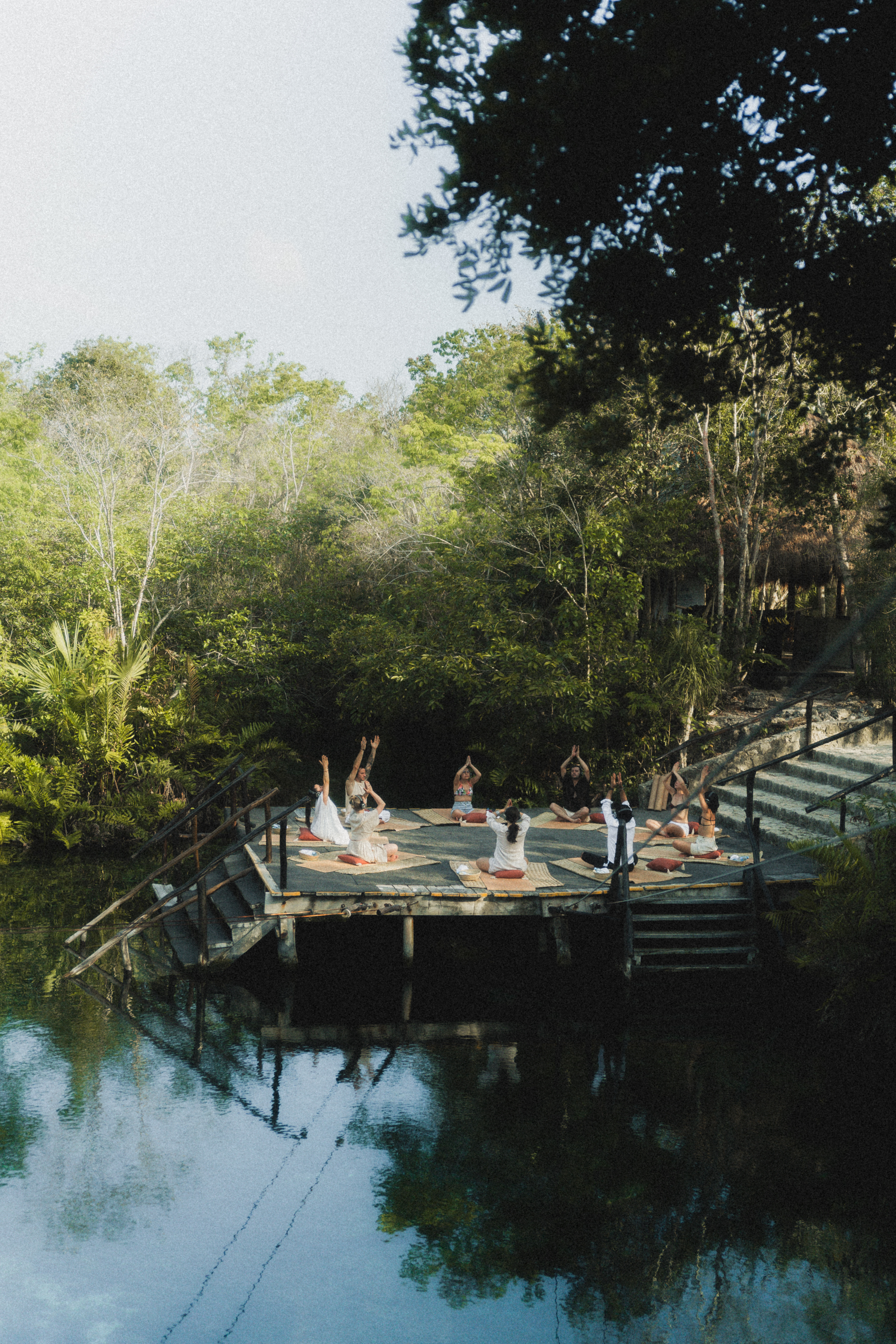 Yoga y meditación grupal en plataforma sobre agua de cenote, rodeado de selva.