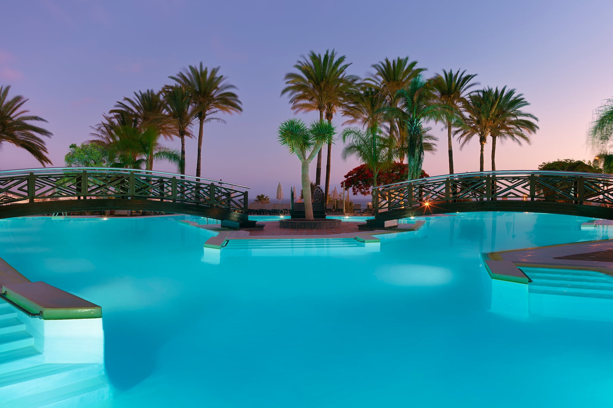 Resort swimming pool with illuminated bridges and palm trees at twilight.