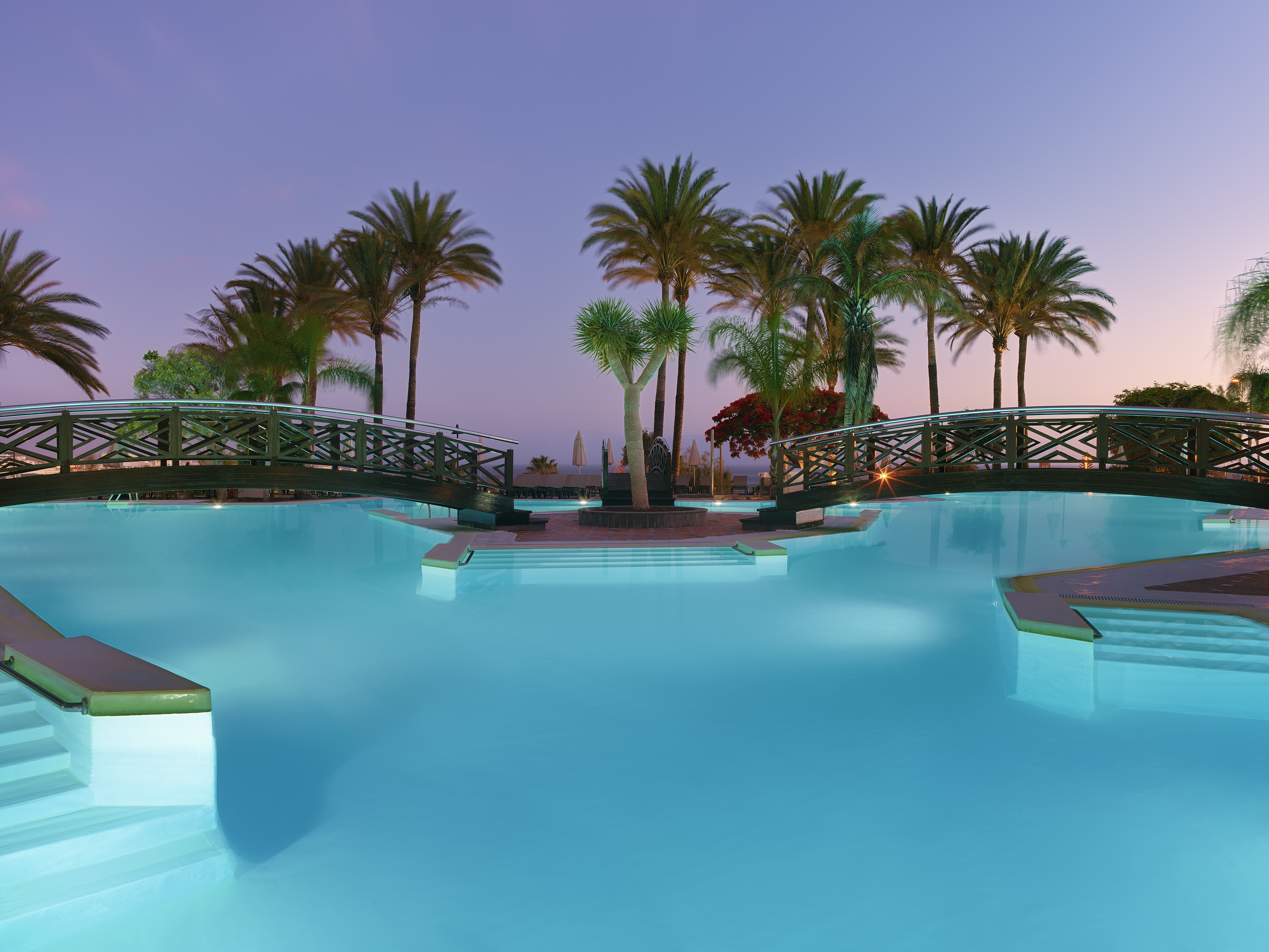 Resort swimming pool with illuminated bridges and palm trees at twilight.