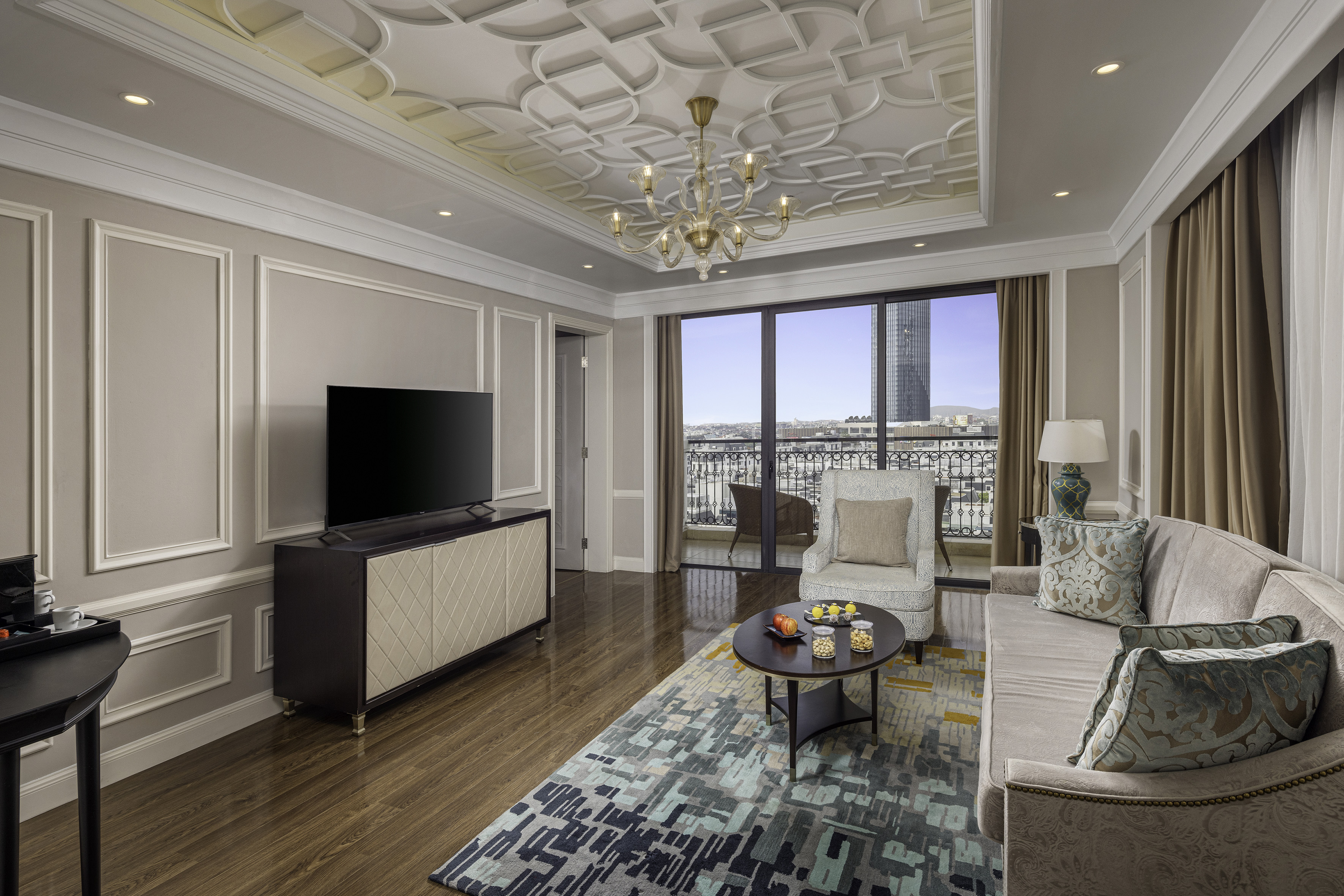 Luxury hotel suite with city view, ornate ceiling, sofa, and TV.