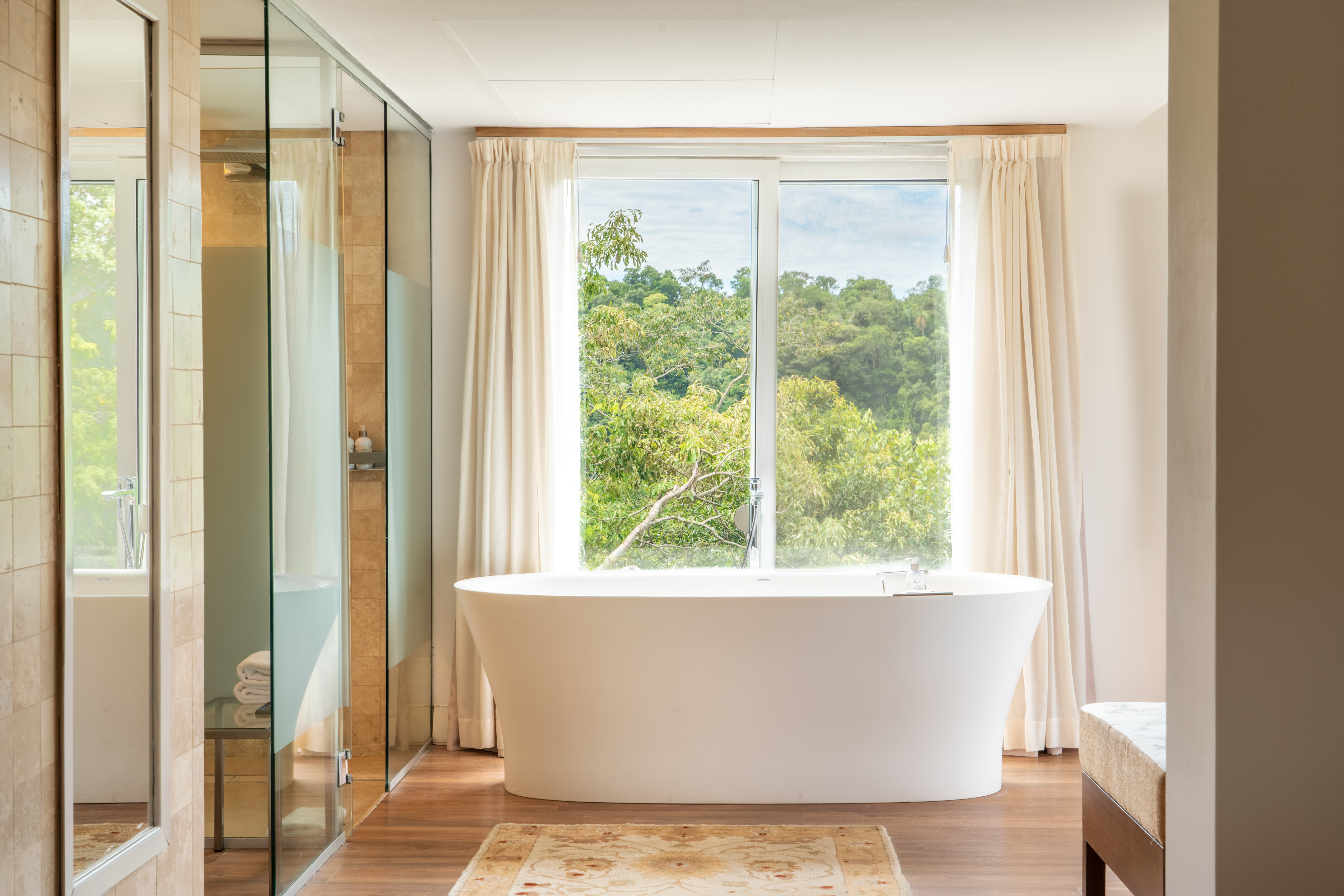 Spa-like bathroom: freestanding tub, tropical forest view, glass shower, wood floor.