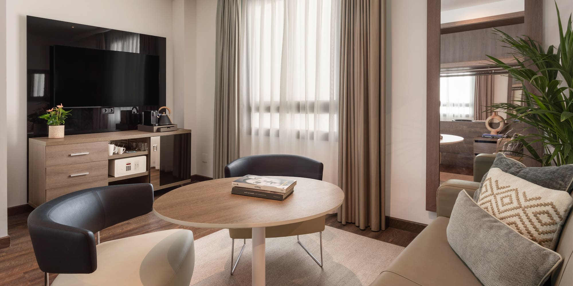 Cozy hotel suite with TV, sitting area, modern furniture, mirror, and bright window.