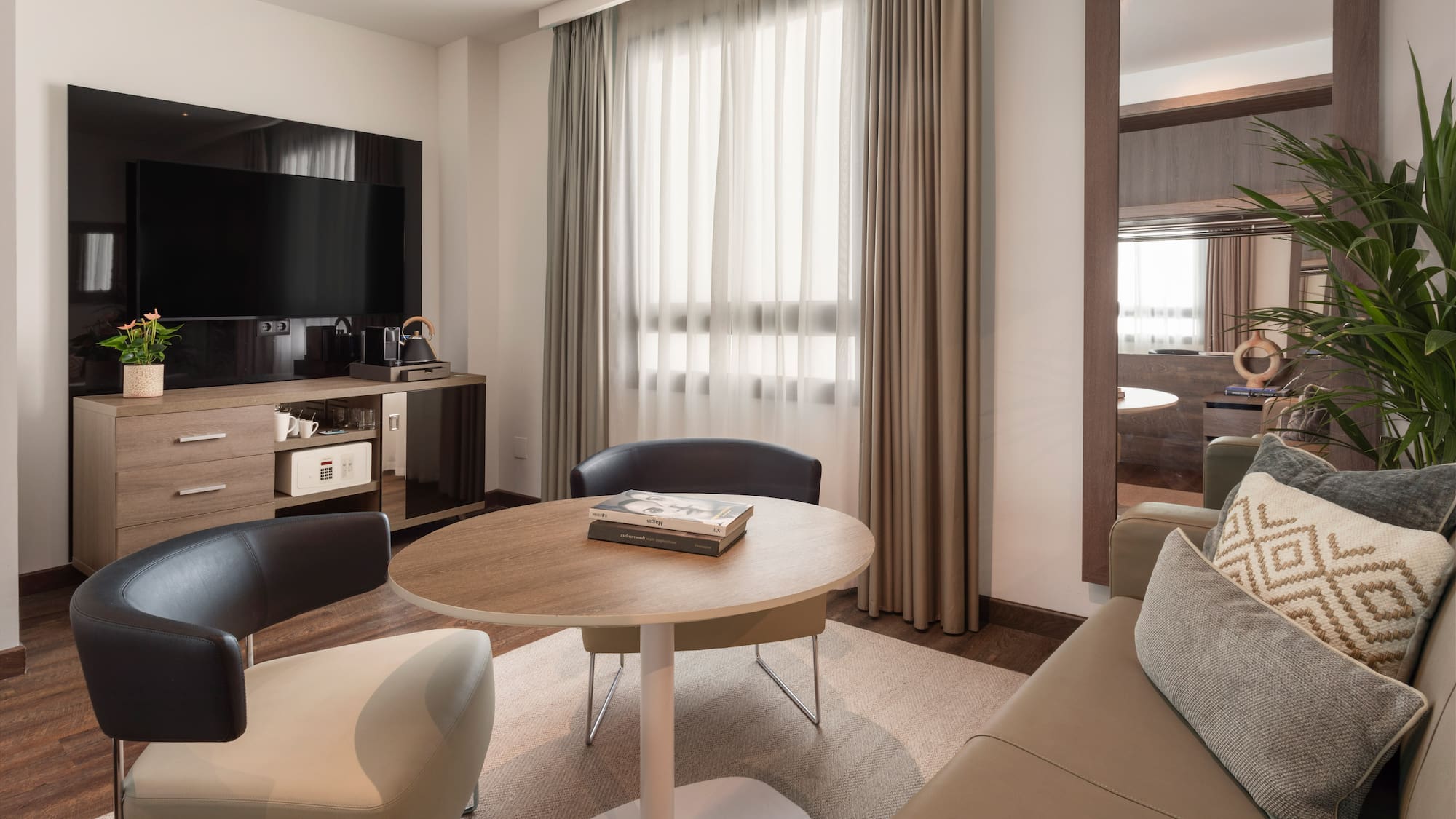 Cozy hotel suite with TV, sitting area, modern furniture, mirror, and bright window.