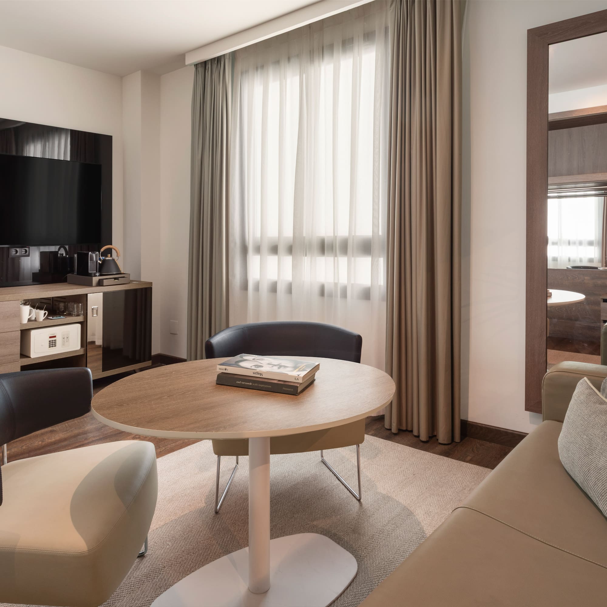 Cozy hotel suite with TV, sitting area, modern furniture, mirror, and bright window.