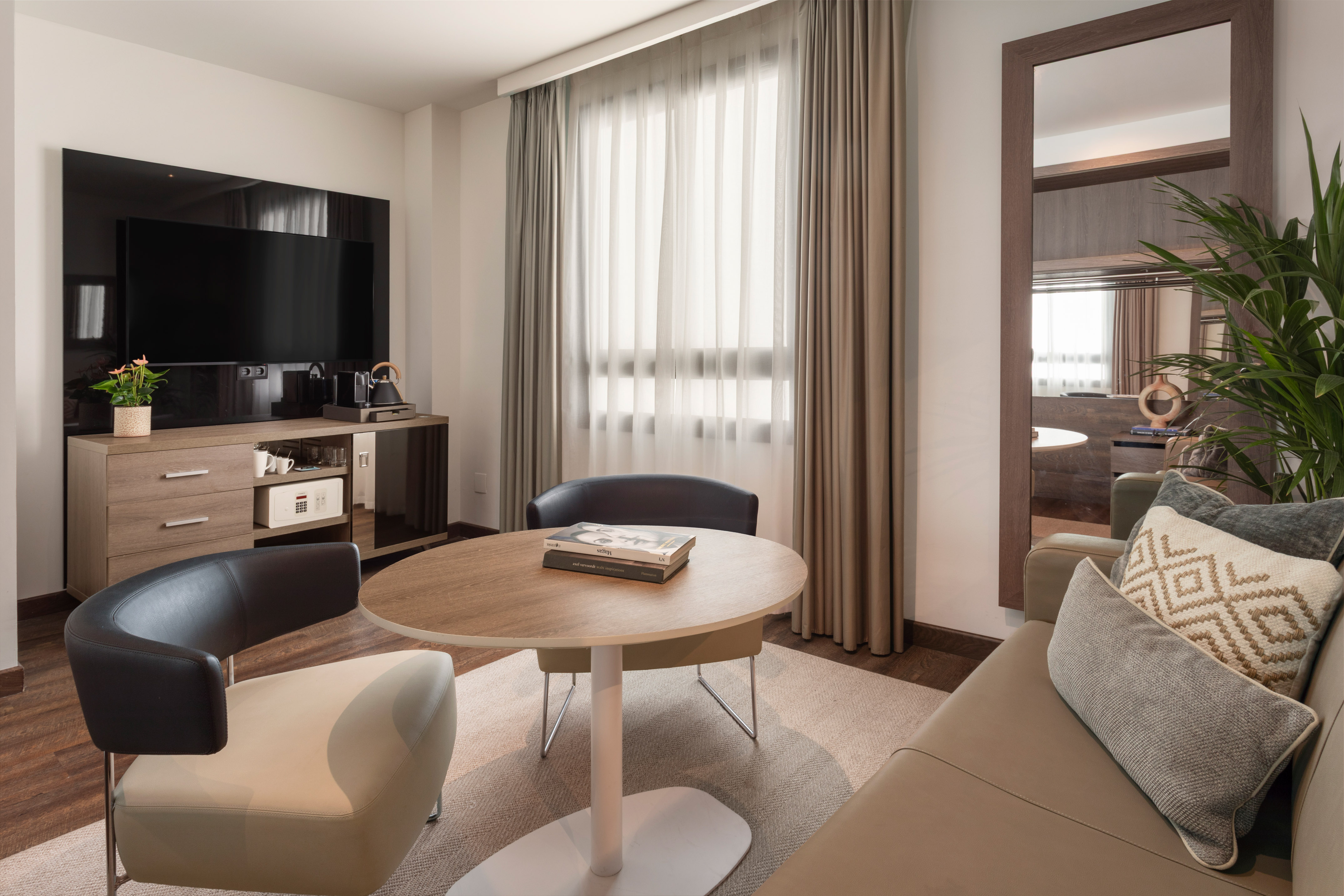 Cozy hotel suite with TV, sitting area, modern furniture, mirror, and bright window.