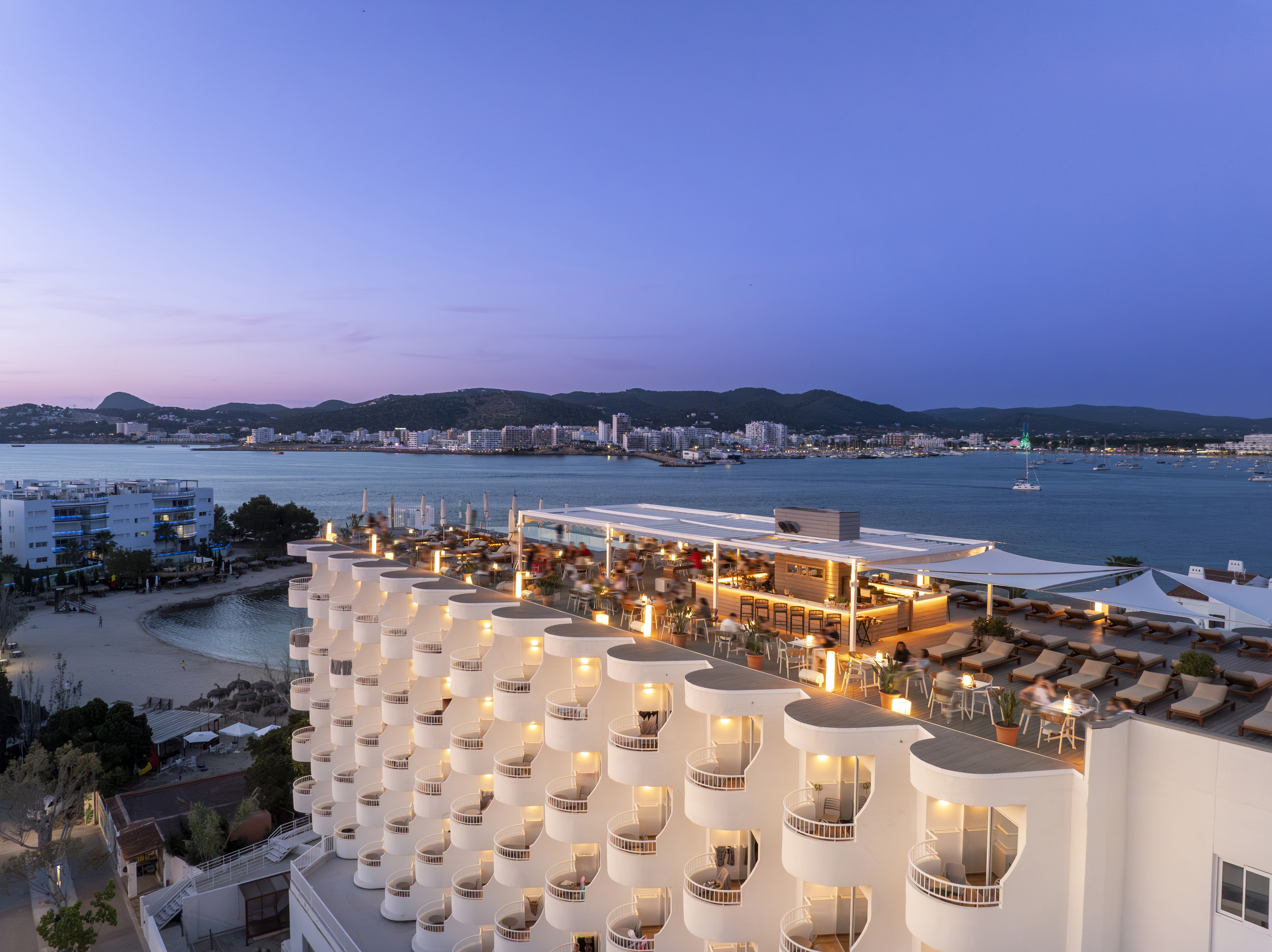 Ibiza hotel rooftop bar with illuminated balconies overlooking the bay at dusk.