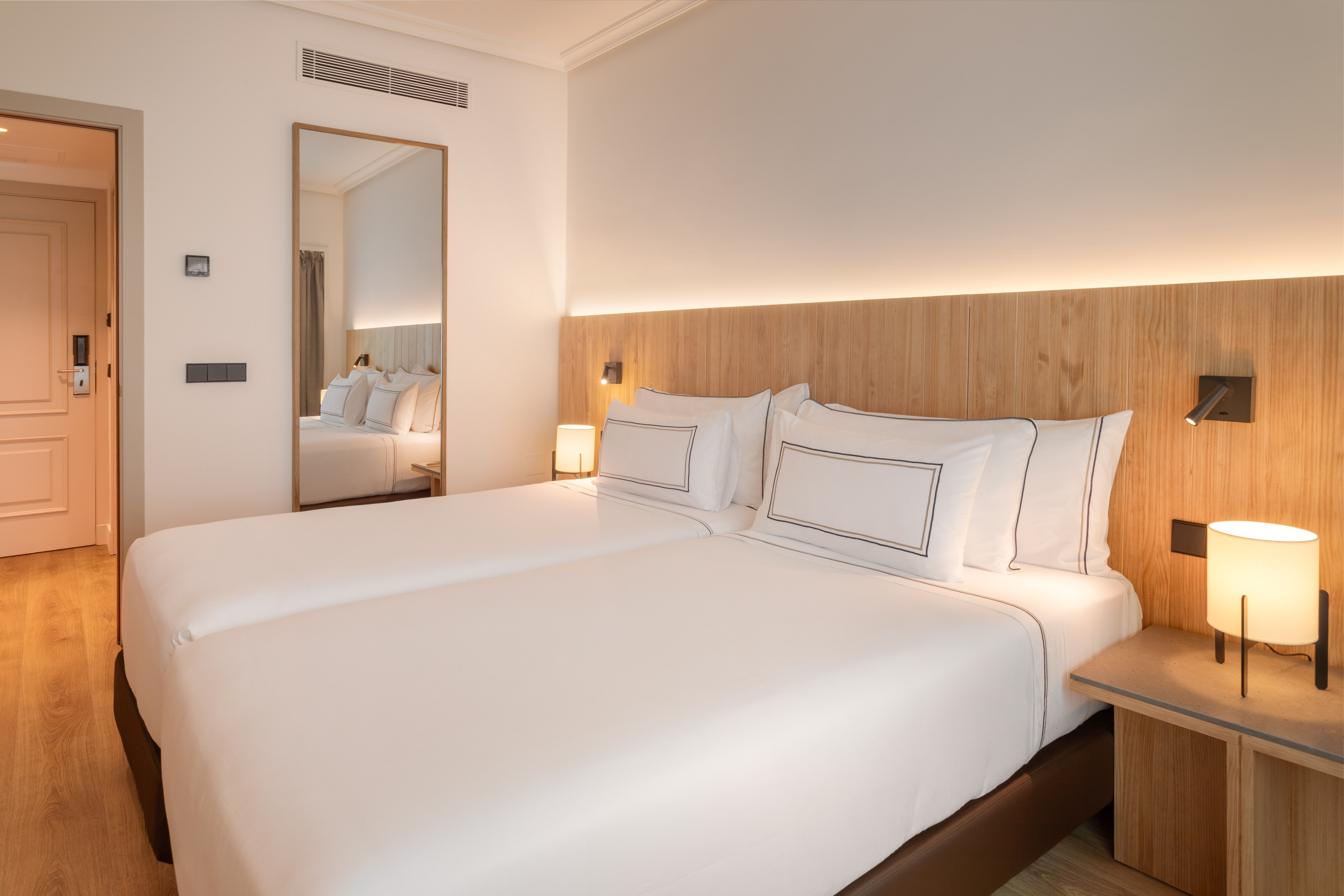 Modern hotel bedroom with twin beds, wooden headboard, and warm ambient lighting.