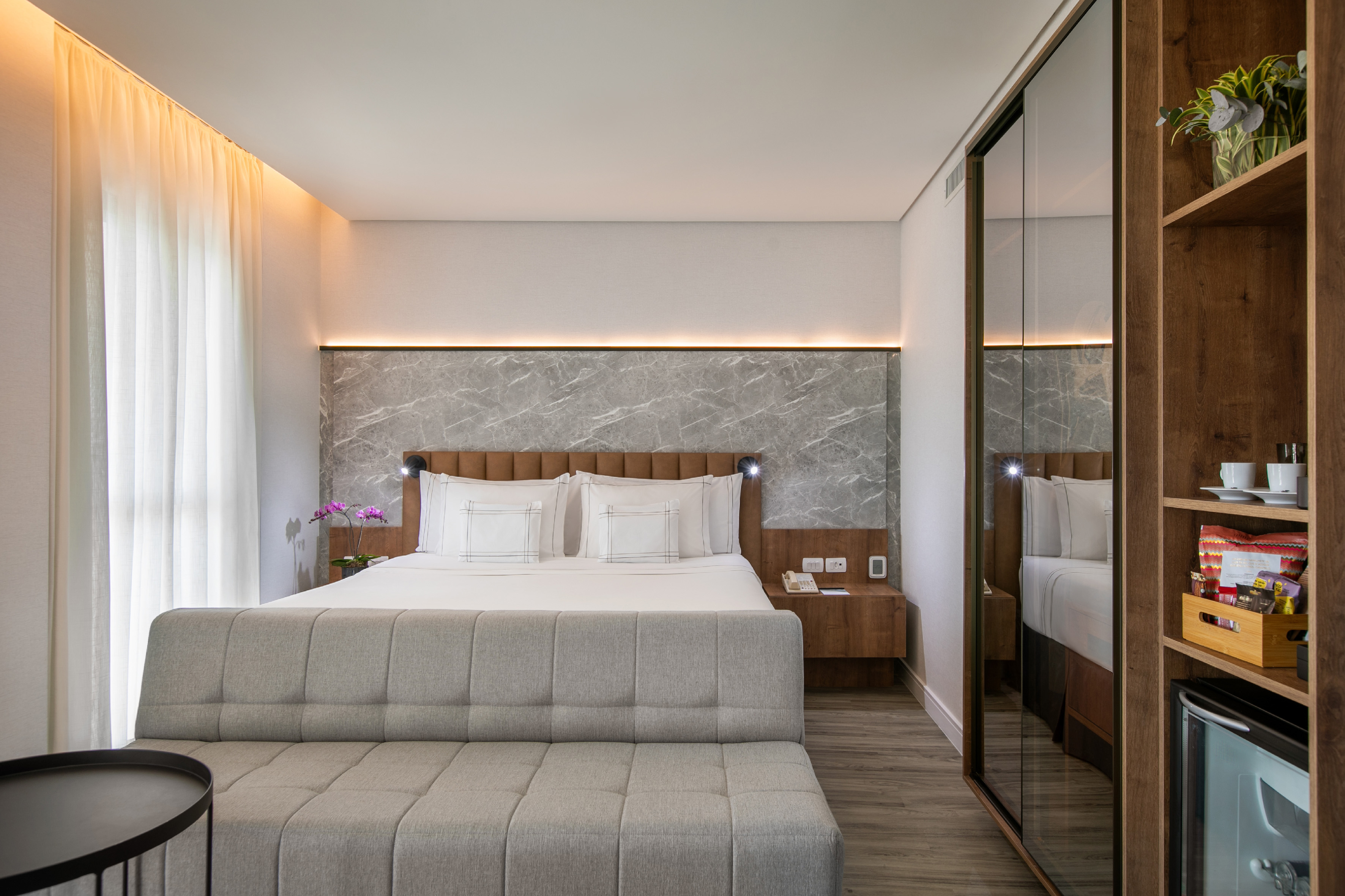 Modern hotel room with large bed, grey accent wall, sofa, and minibar.