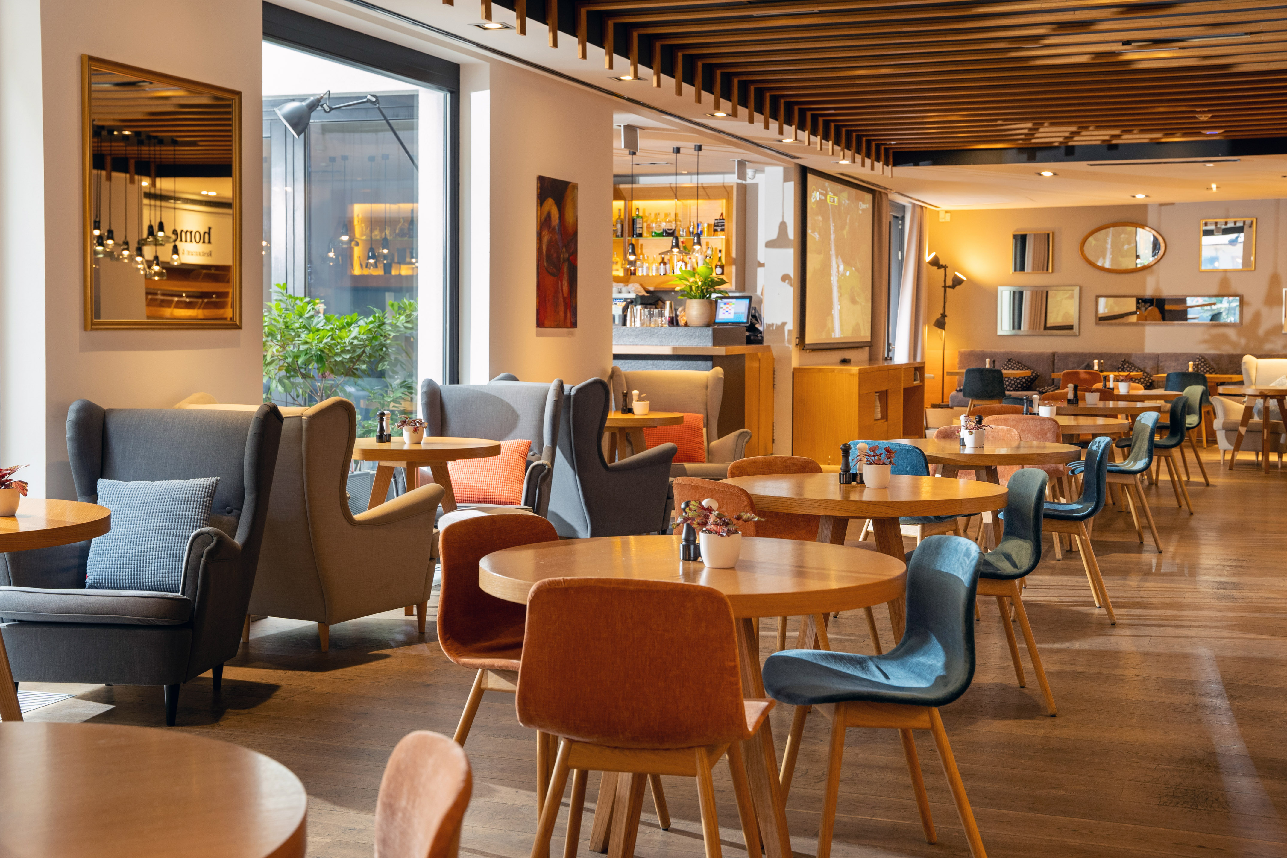 Modern restaurant dining room with wooden tables, varied chairs, and bar area.