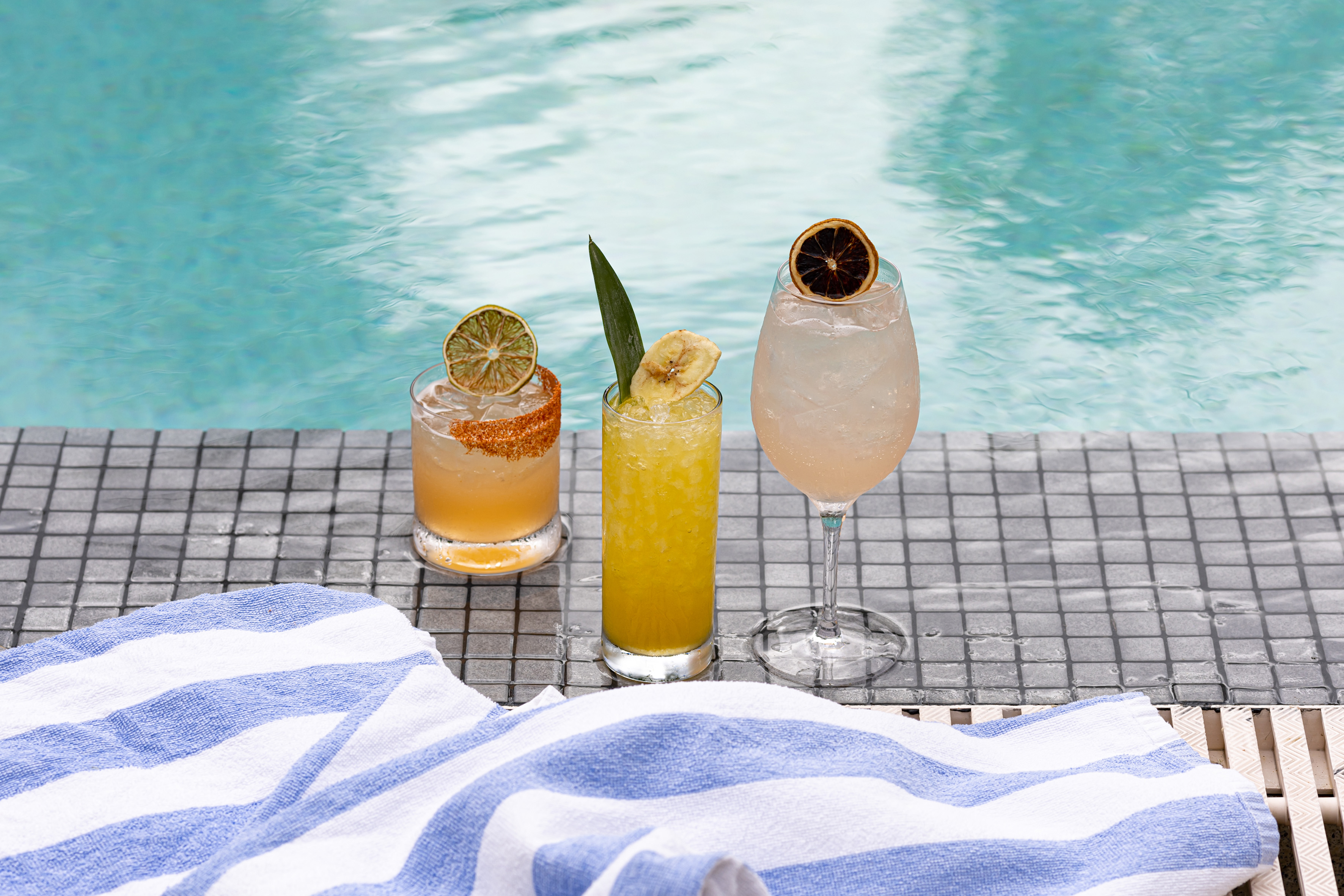 Tropical poolside cocktails with fruit and towel by a blue summer pool.