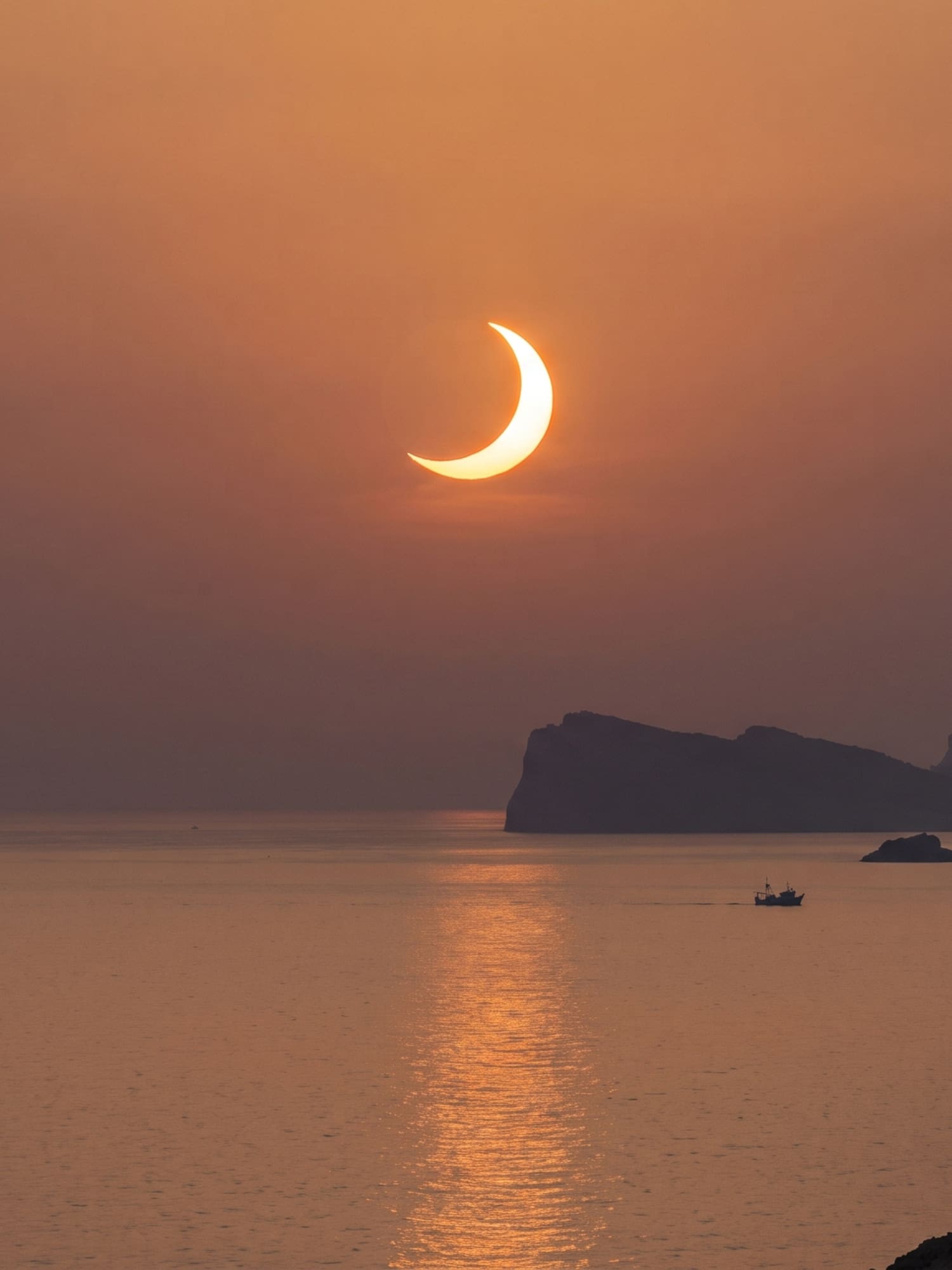 Sol creciente de eclipse sobre el mar al atardecer, con montañas y barco.