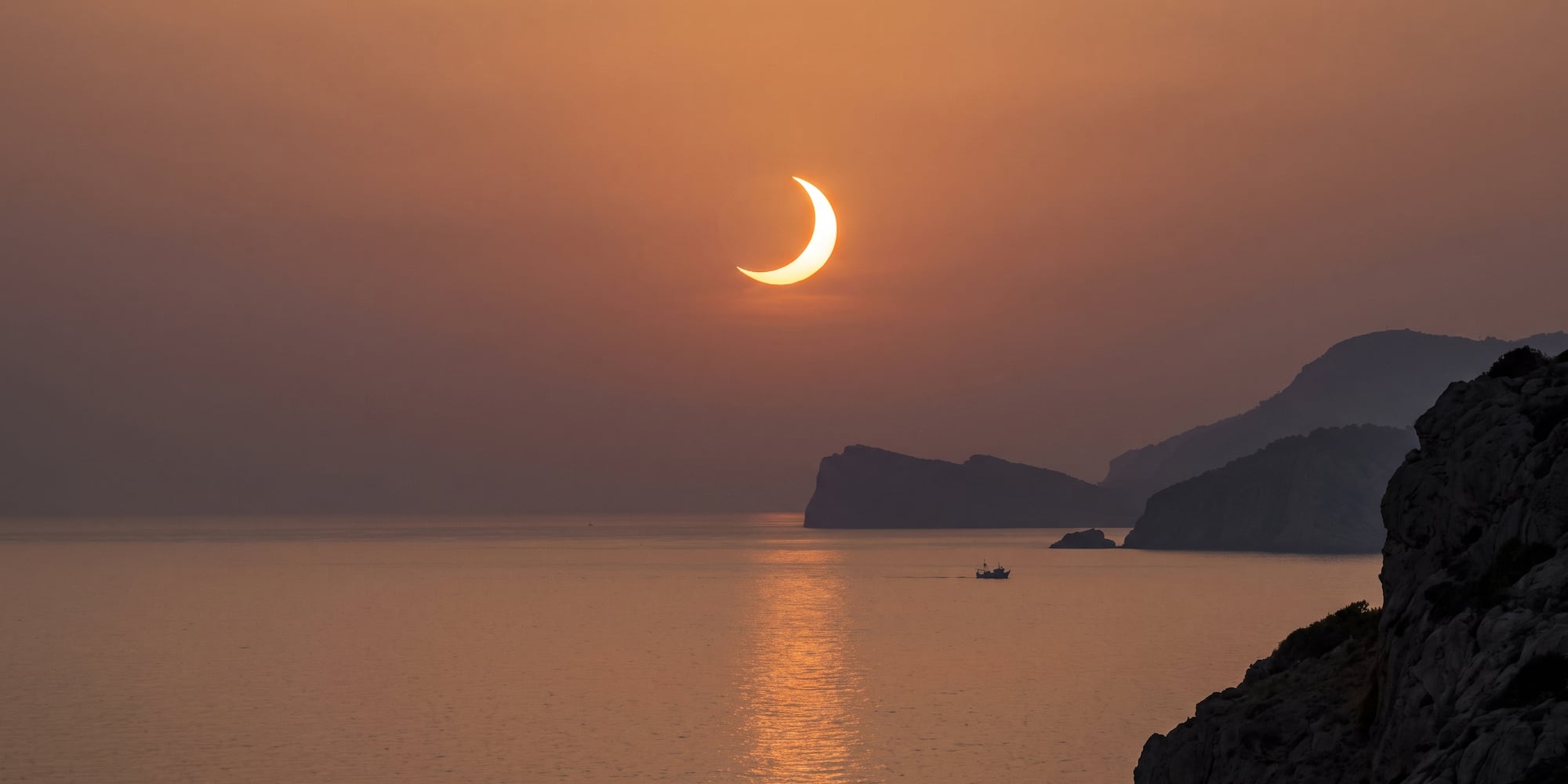 Eclipse solar de media luna sobre el mar, montañas y barco al atardecer.