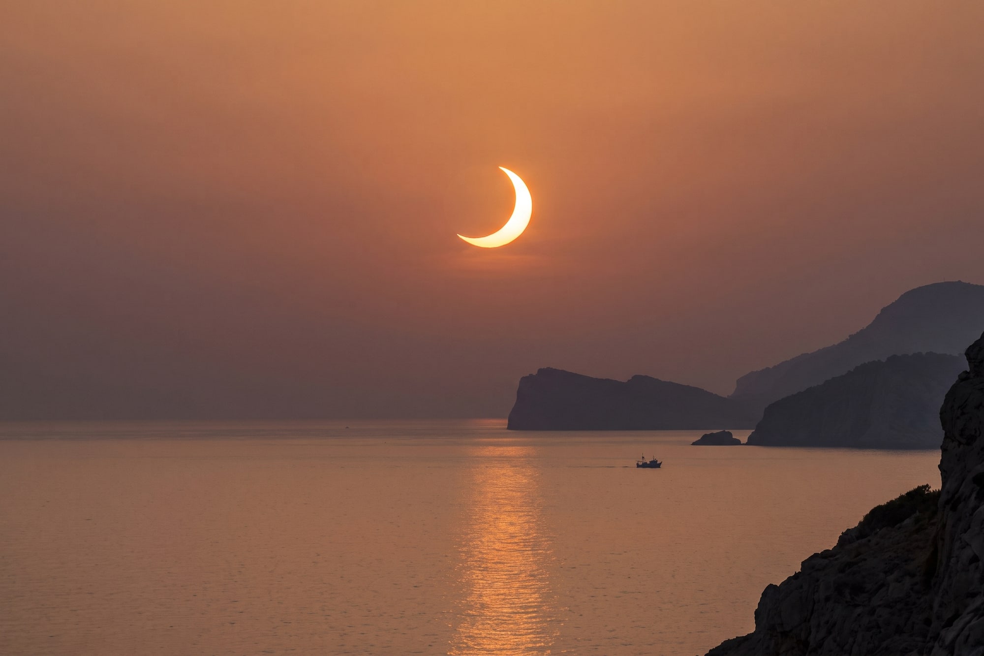 Sol creciente de eclipse sobre el mar al atardecer, con montañas y barco.