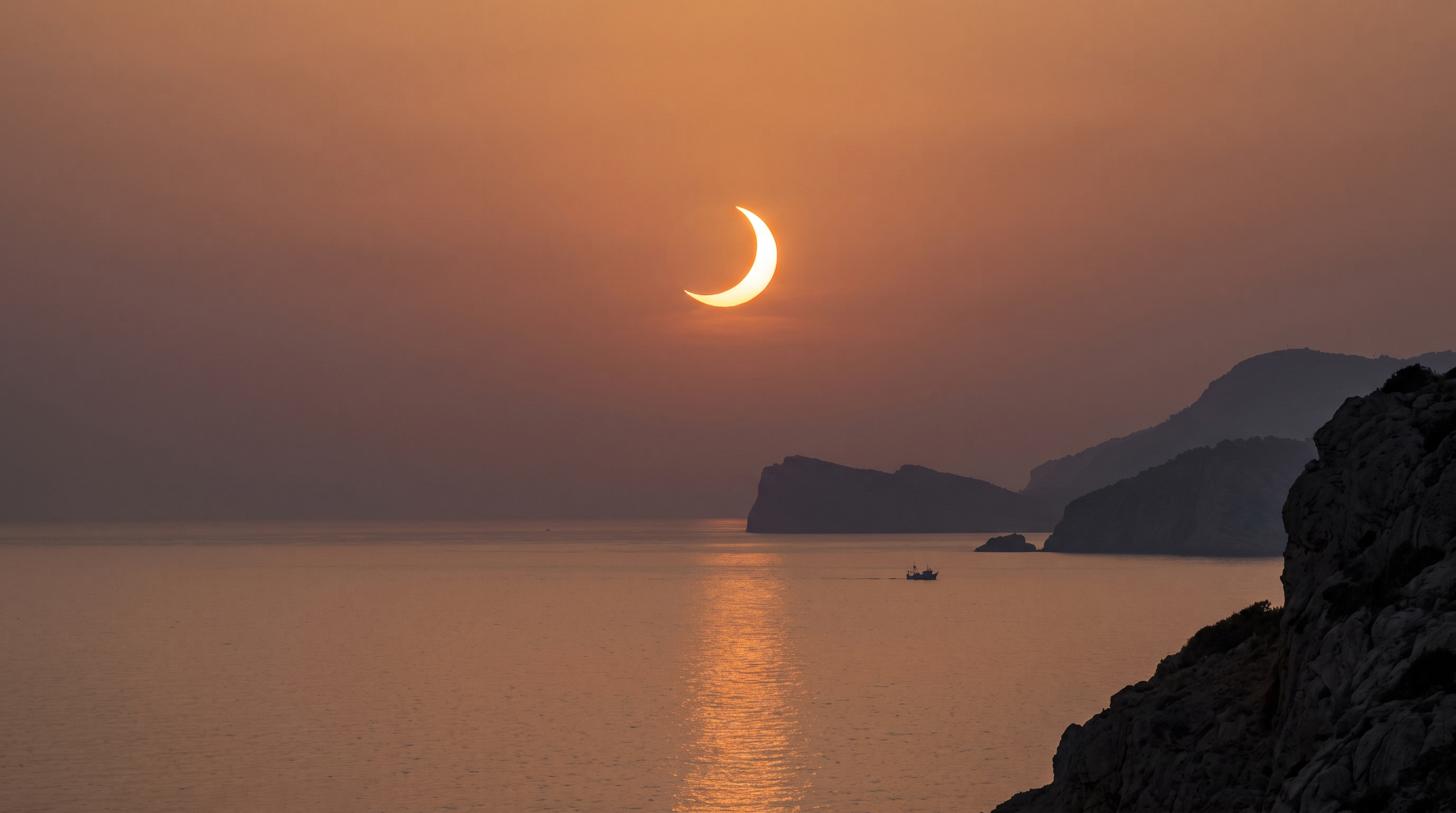 Crescent sun solar eclipse over ocean, silhouetted mountains and boat.