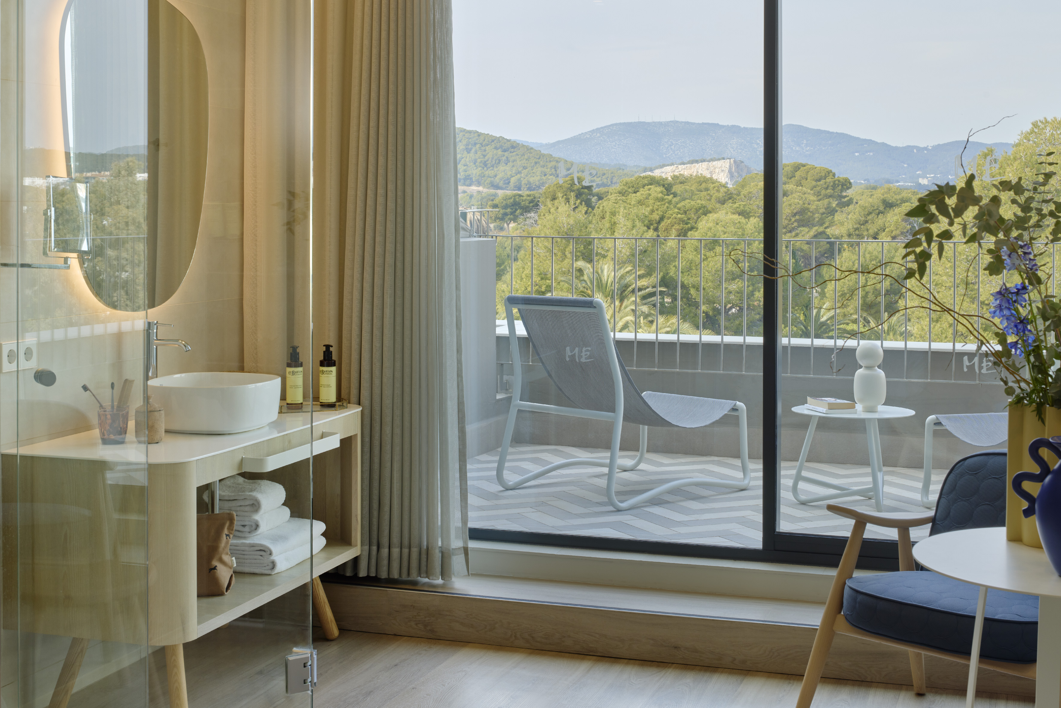 Hotel bathroom vanity, balcony lounge chair, lush green mountain view.