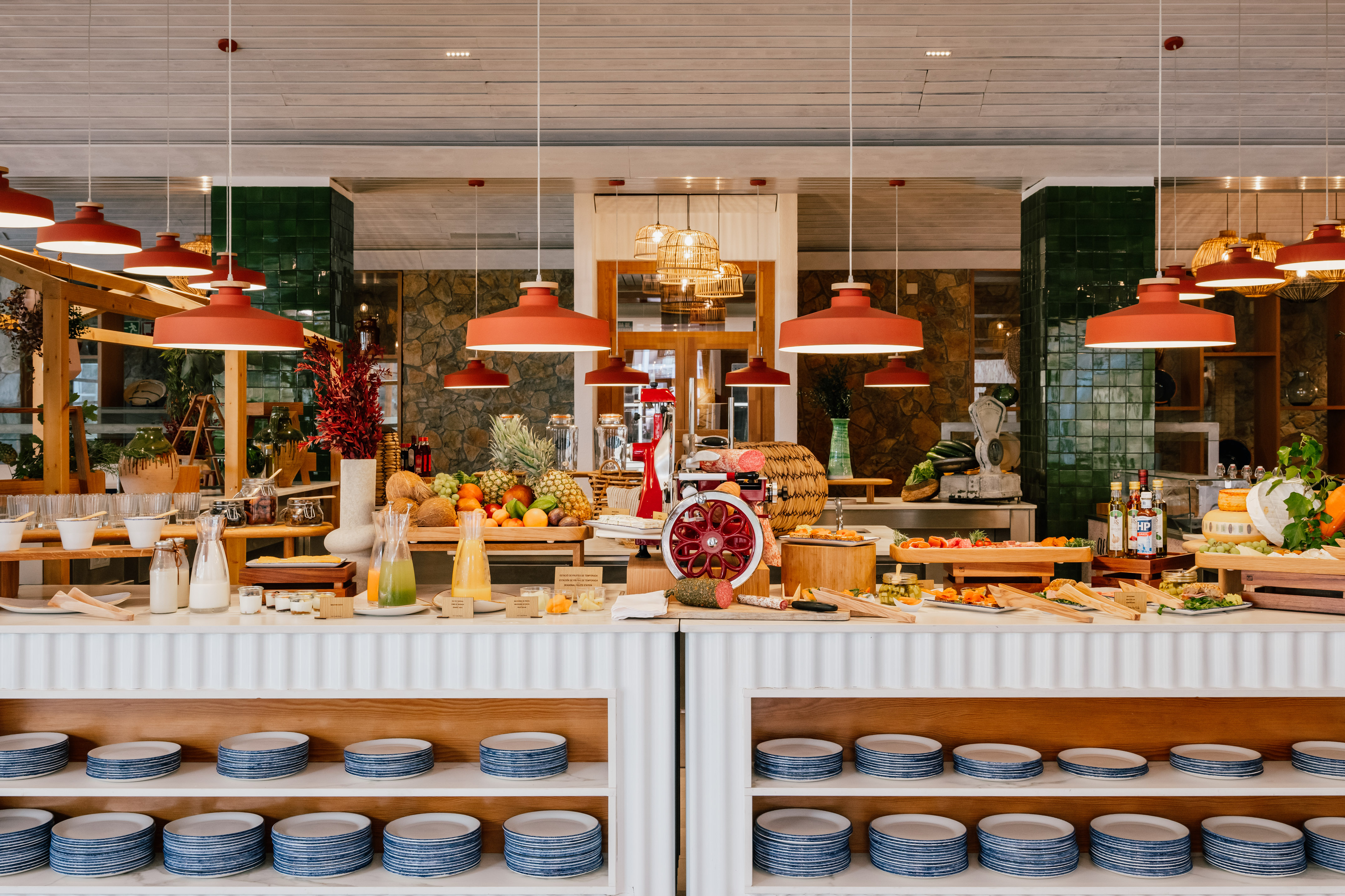 Hotel buffet with fresh fruits, cured meats, cheeses, juices, and stacked plates.