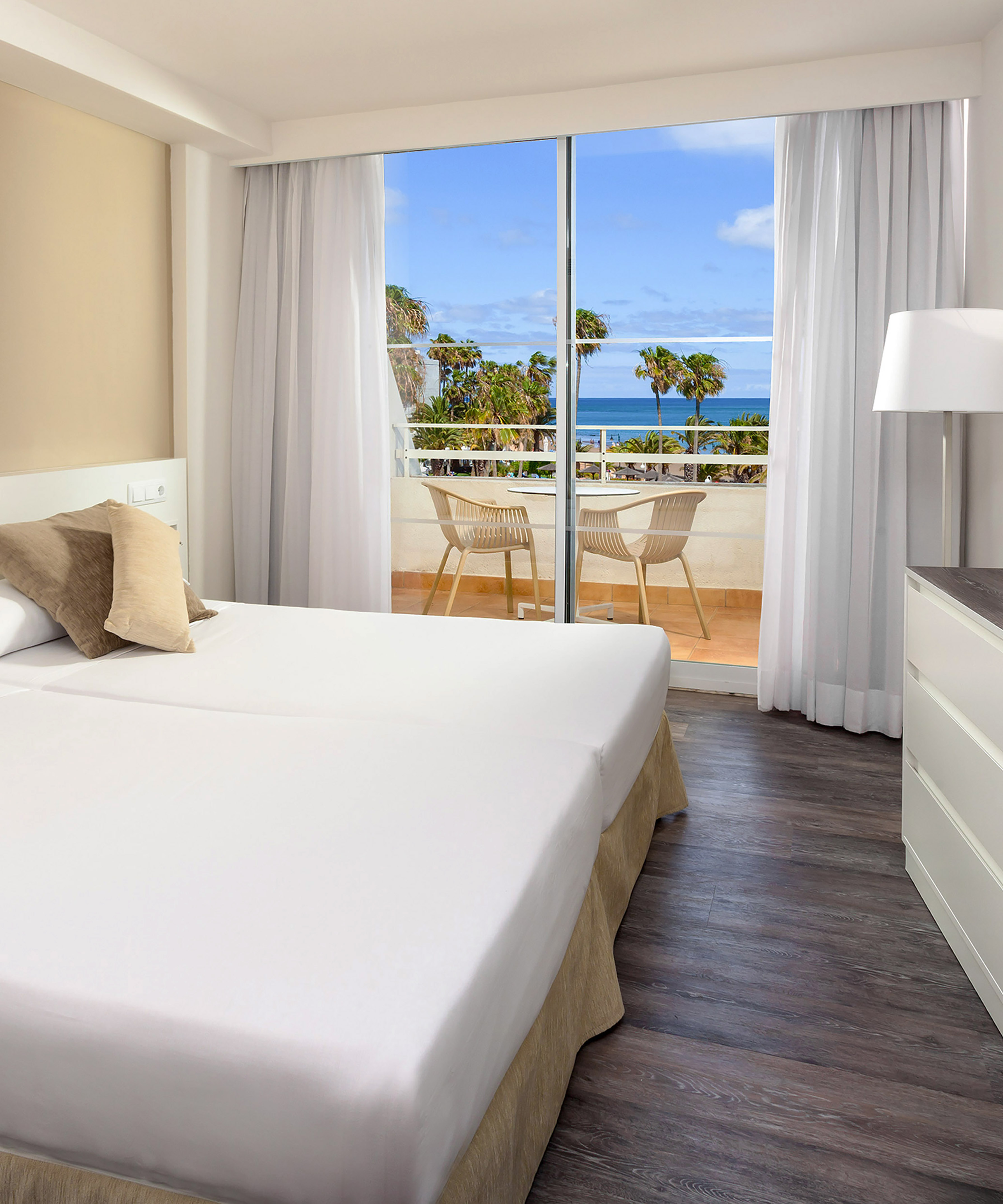 Bright hotel room with twin beds, ocean view balcony, and palm trees.
