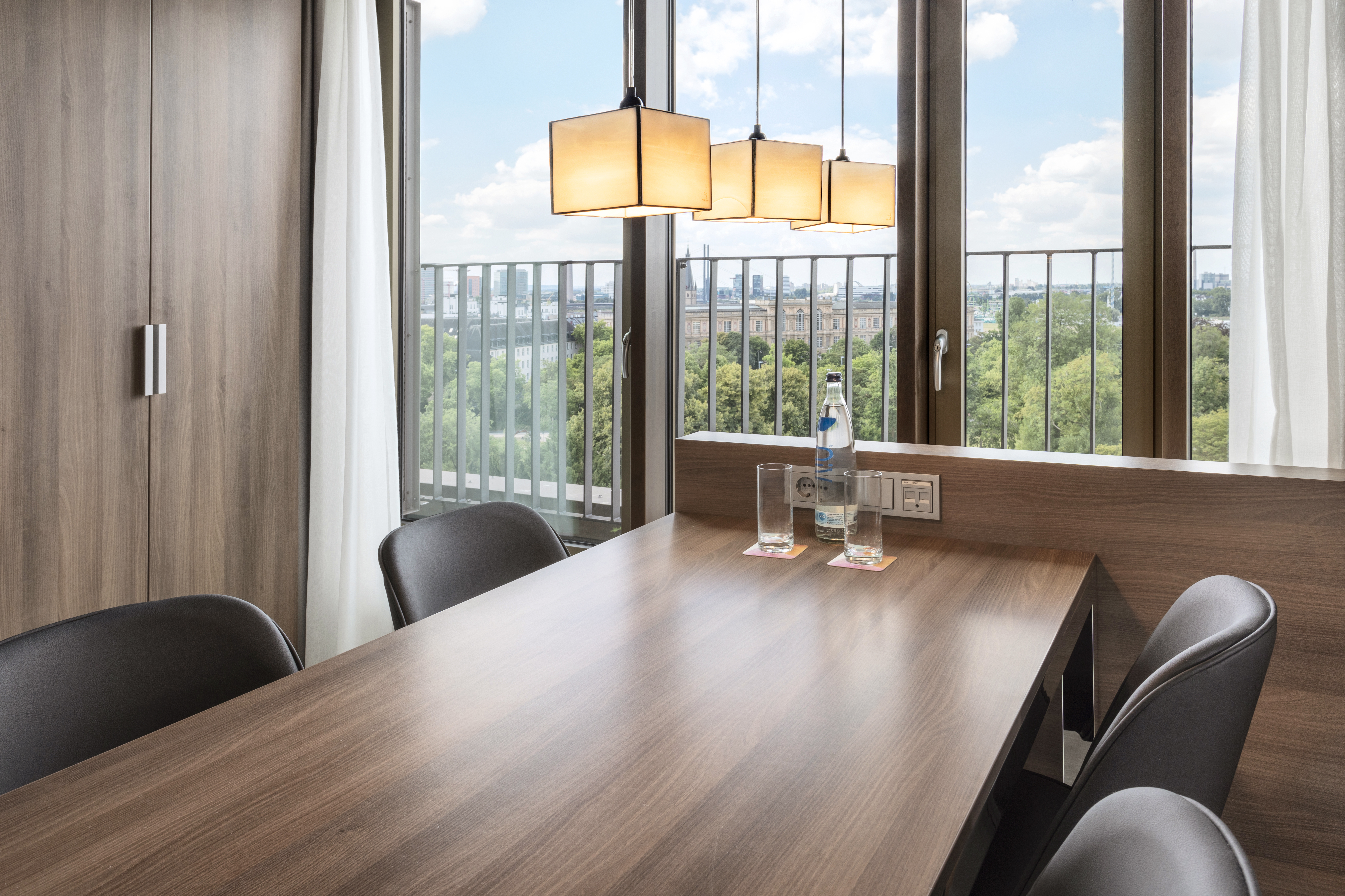 Modern hotel room with wooden table, chairs, city view, and pendant lights.