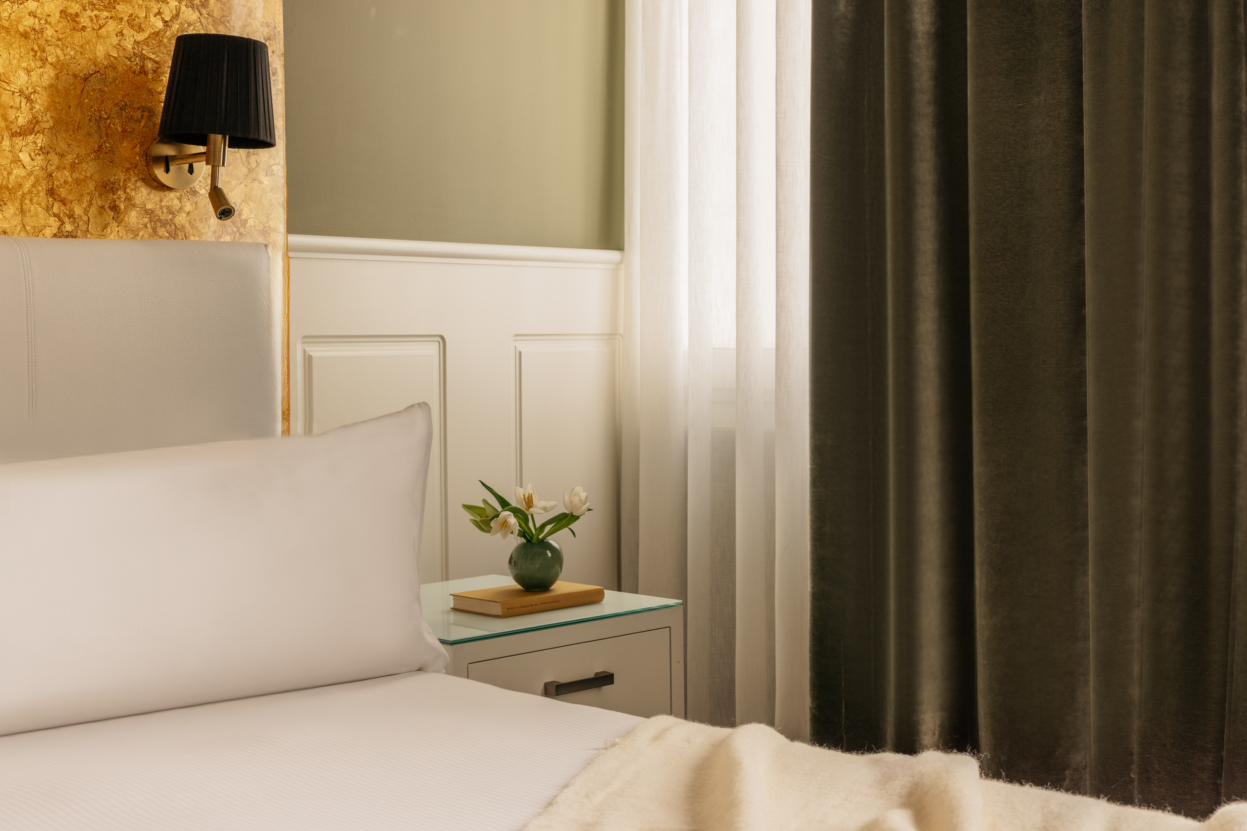 Dormitorio de hotel con cama, pared dorada, cabecero blanco y escritorio.