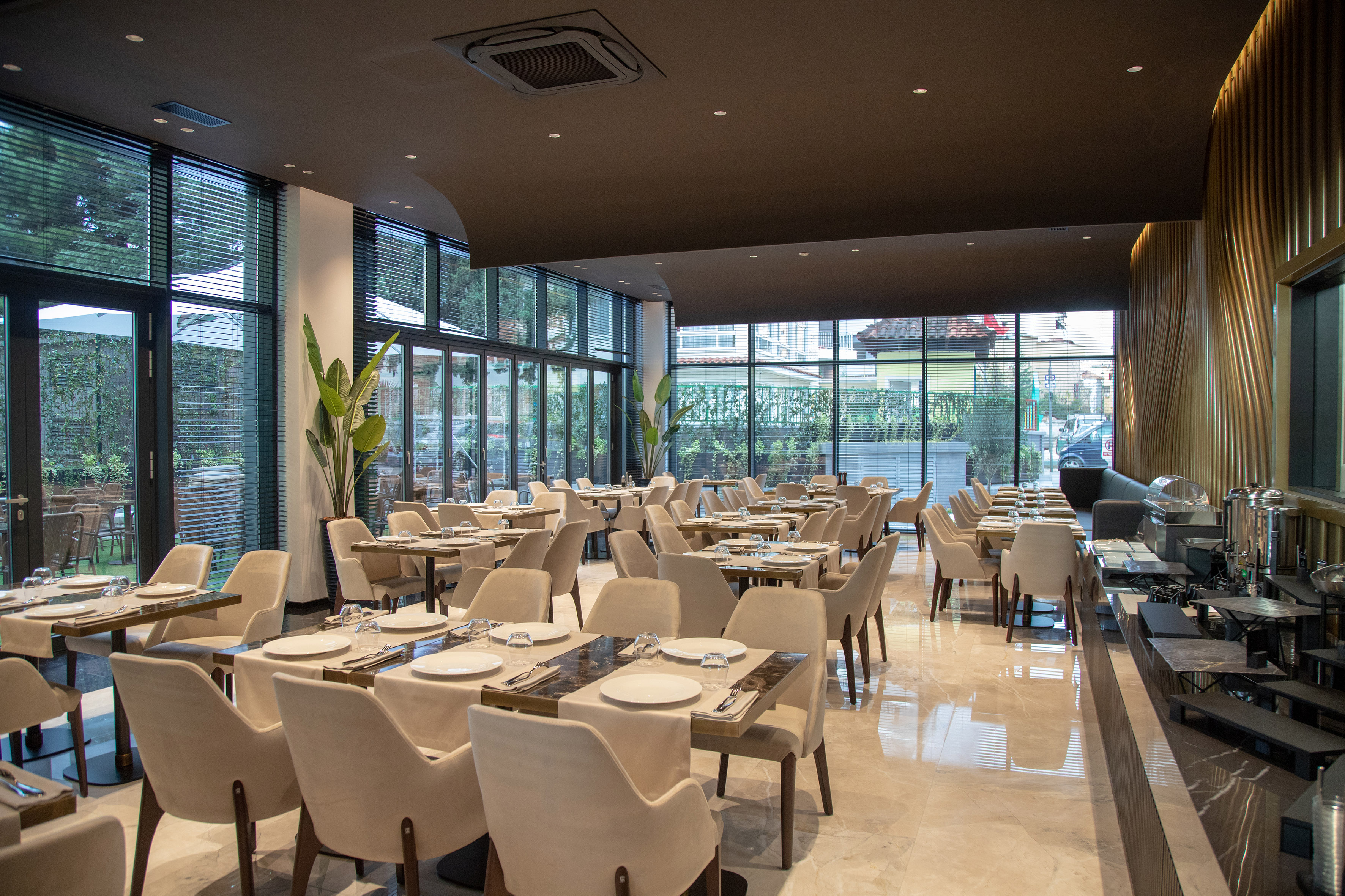 Modern restaurant dining room with tables, beige chairs, large windows, buffet.