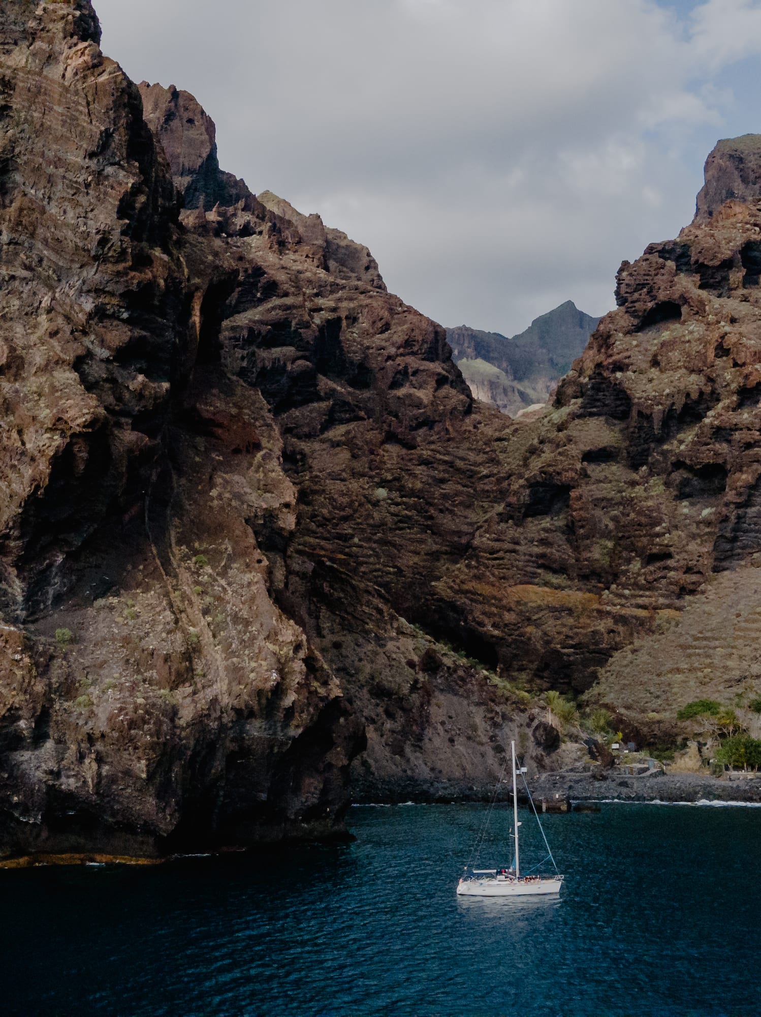 White sailboat on blue water, framed by towering volcanic cliffs.