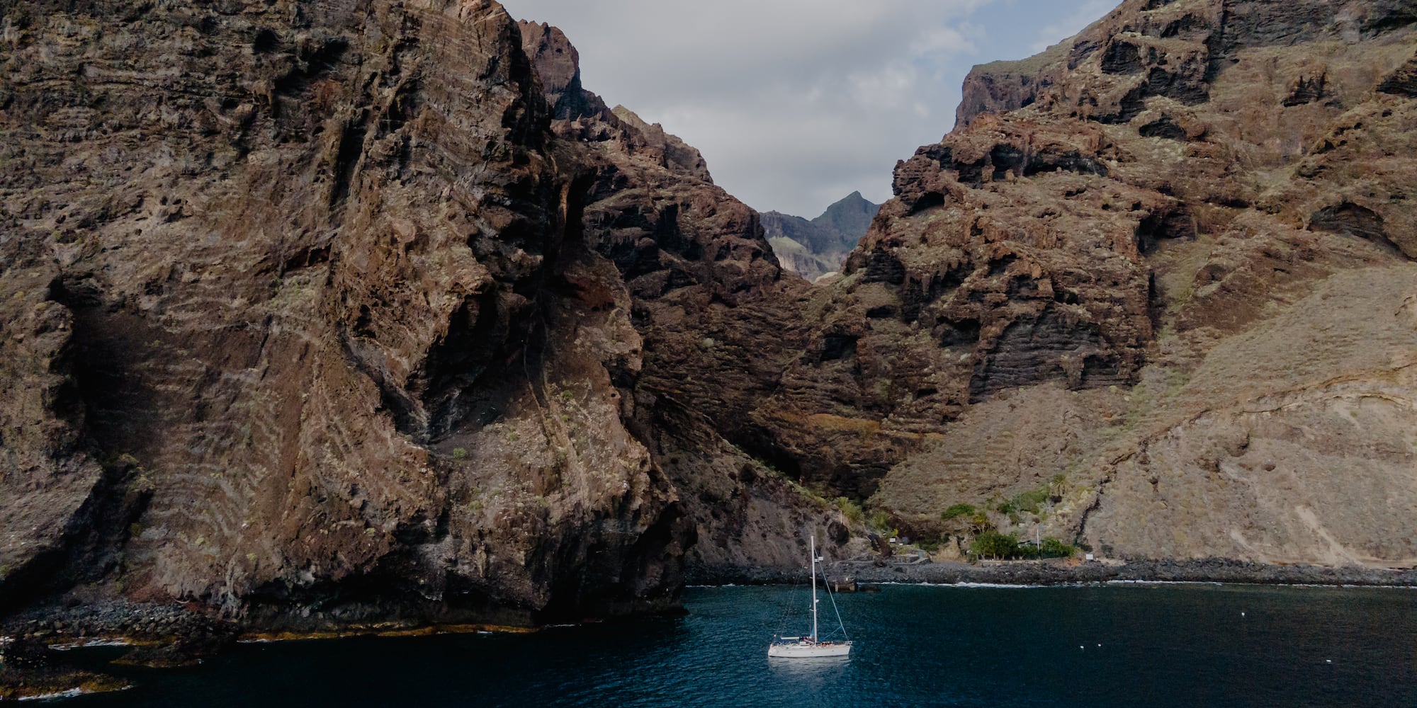 White sailboat on blue water, framed by towering volcanic cliffs.