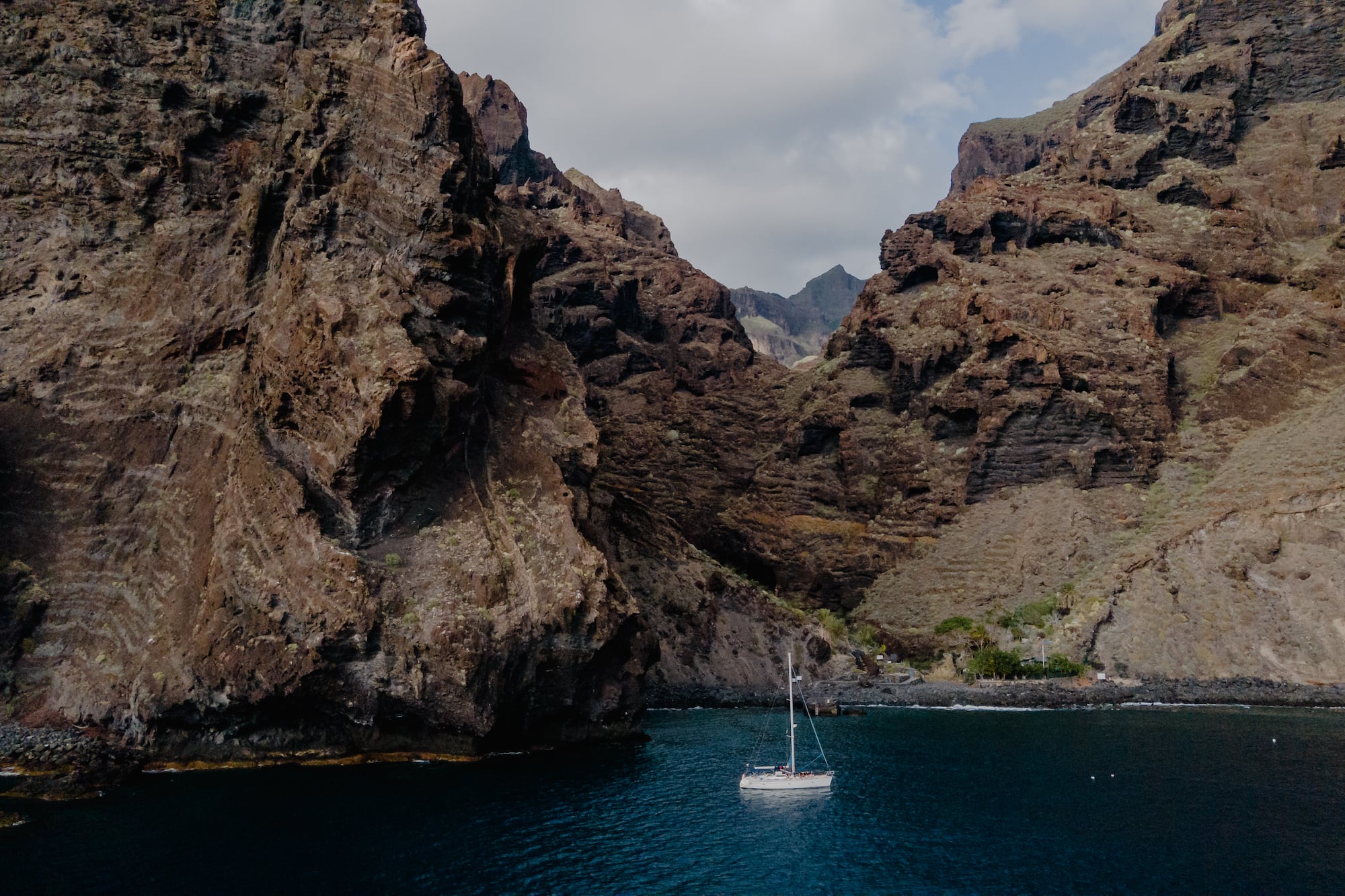 White sailboat on blue water, framed by towering volcanic cliffs.