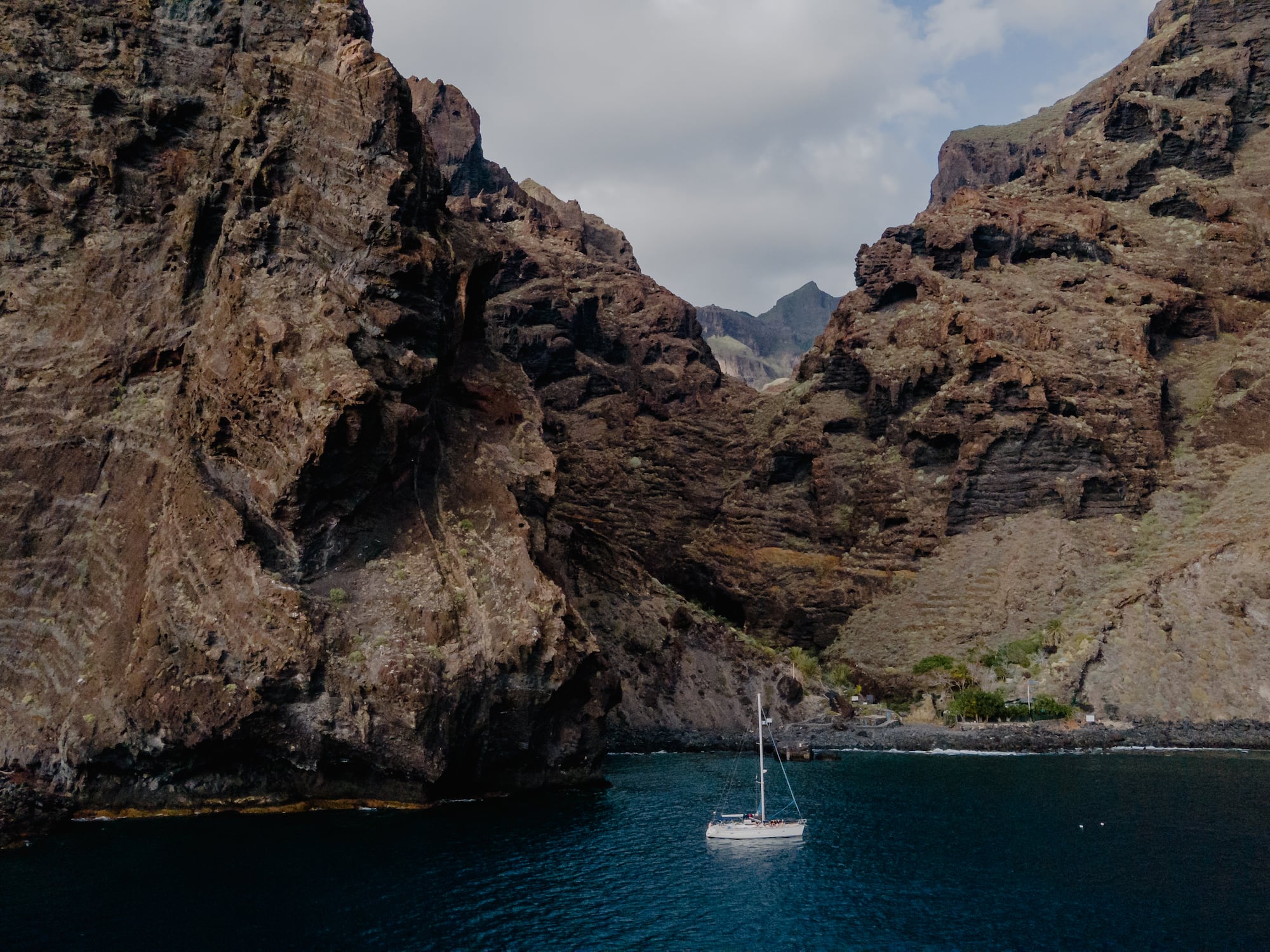 White sailboat on blue water, framed by towering volcanic cliffs.
