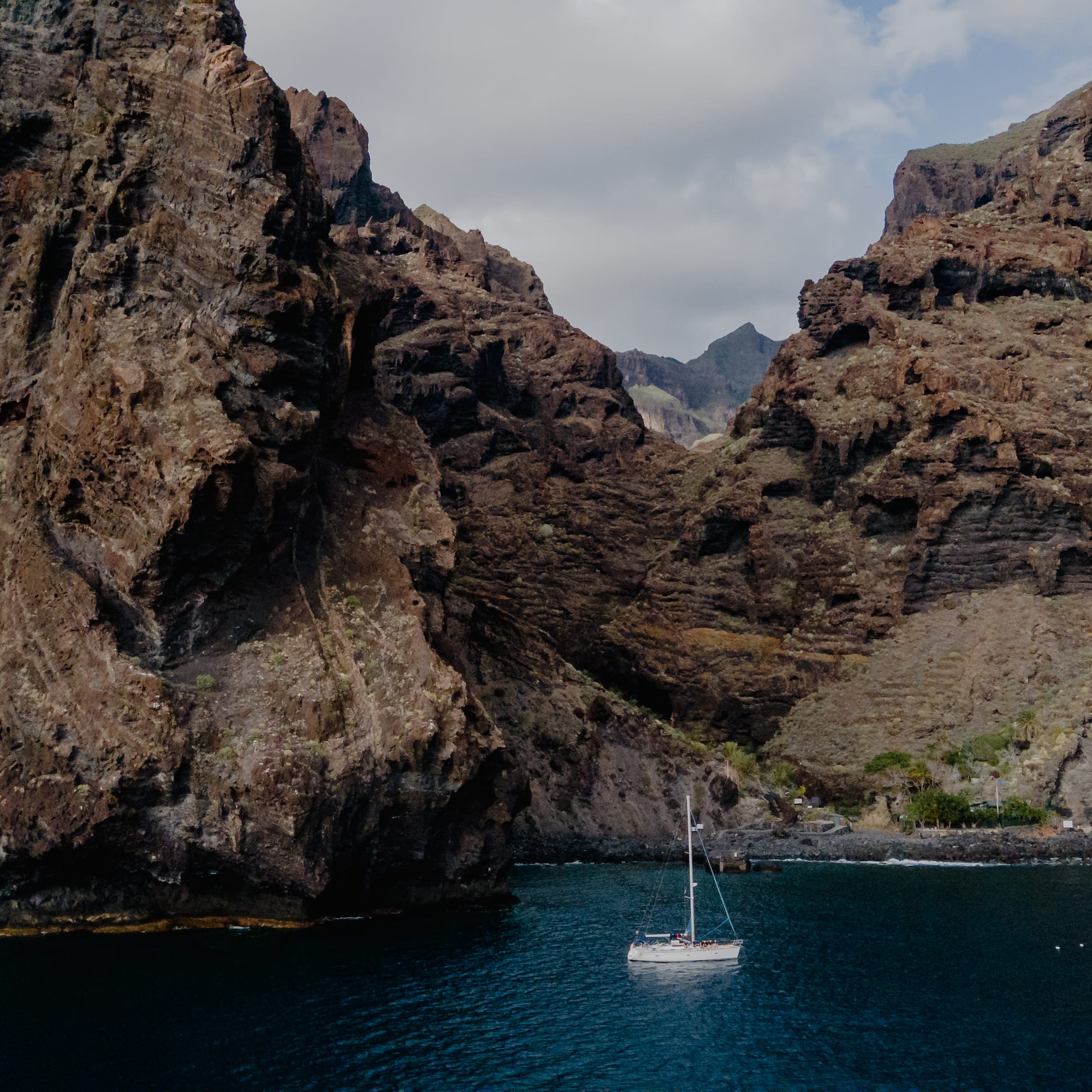 White sailboat on blue water, framed by towering volcanic cliffs.