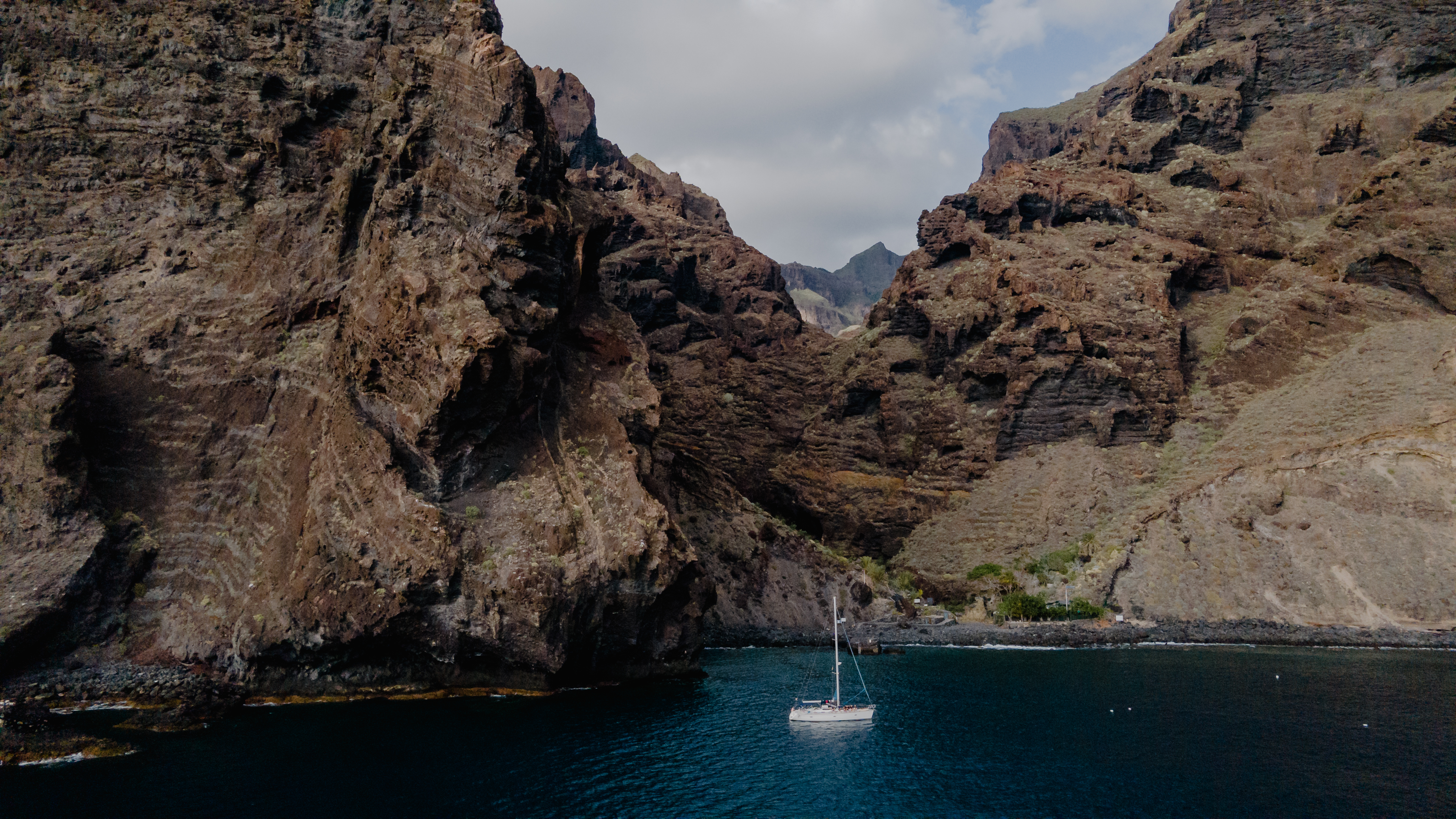White sailboat on blue water, framed by towering volcanic cliffs.