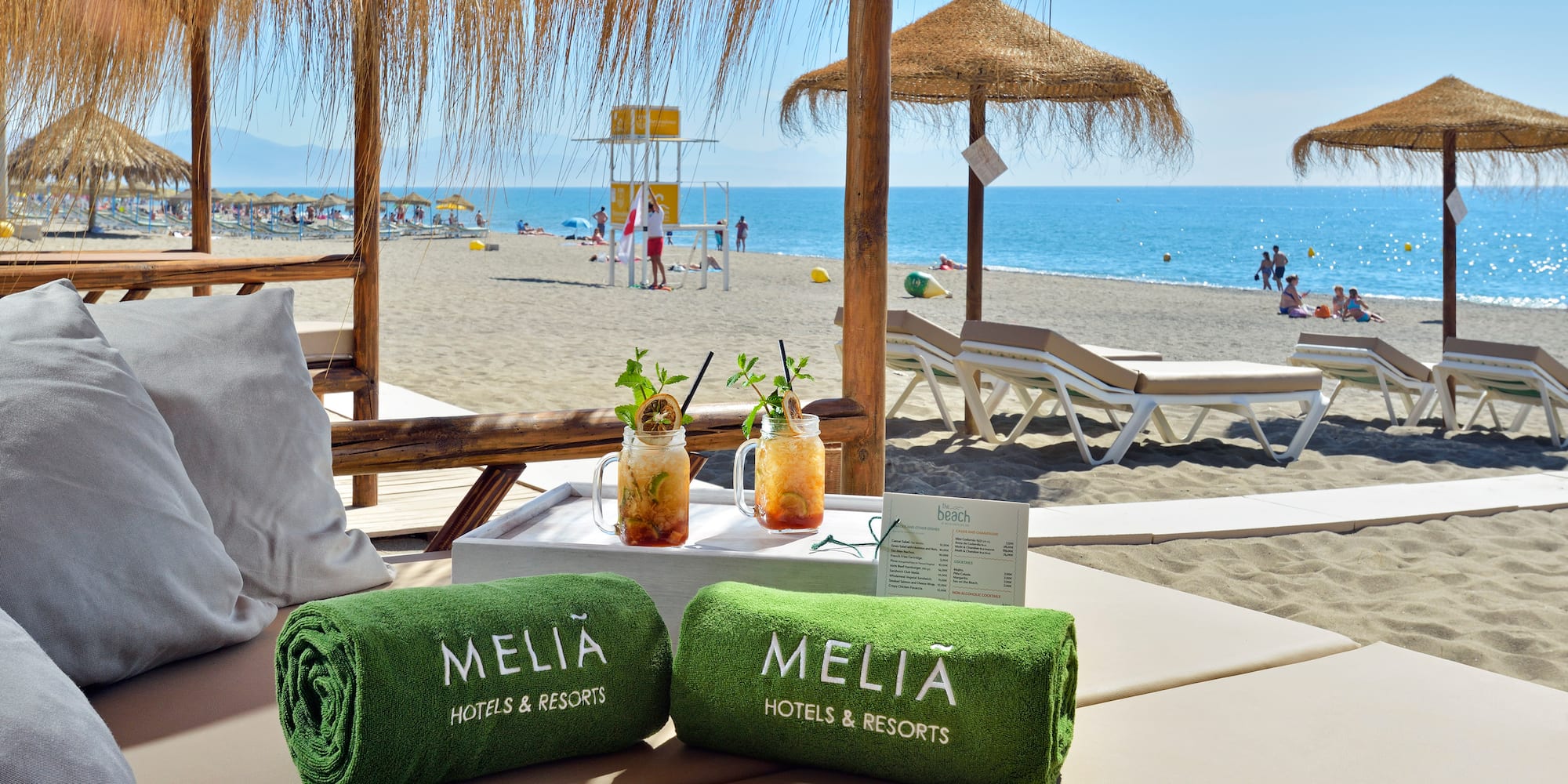 Meliá branded towels, tropical cocktails on beach daybed by ocean, umbrellas.