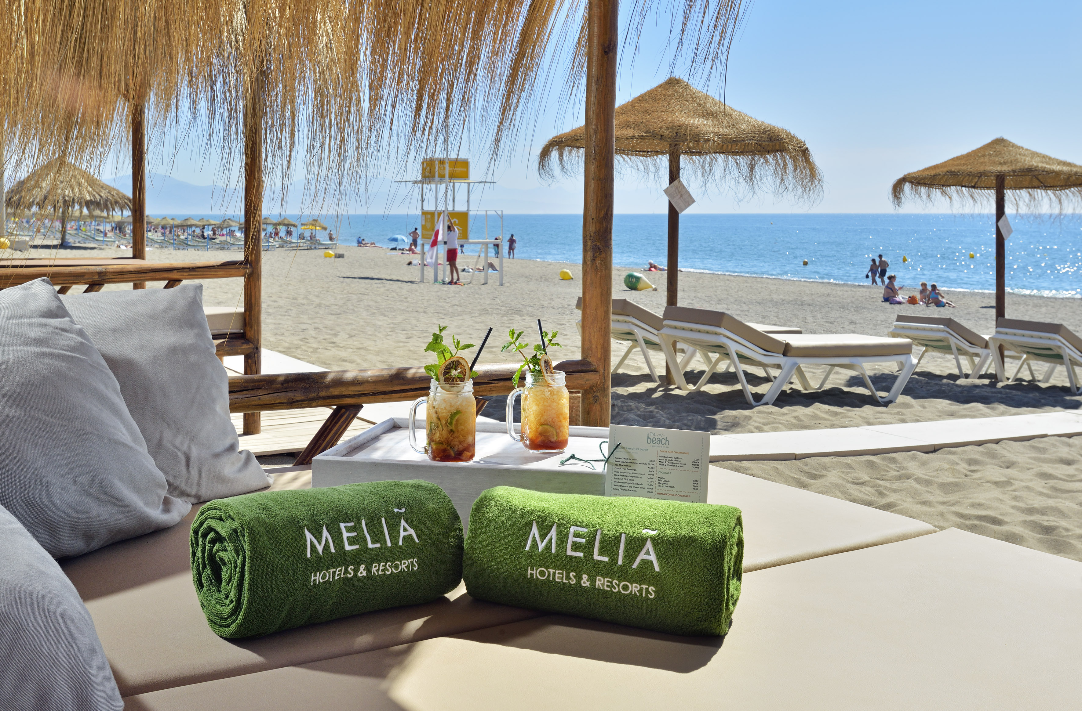 Meliá branded towels, tropical cocktails on beach daybed by ocean, umbrellas.