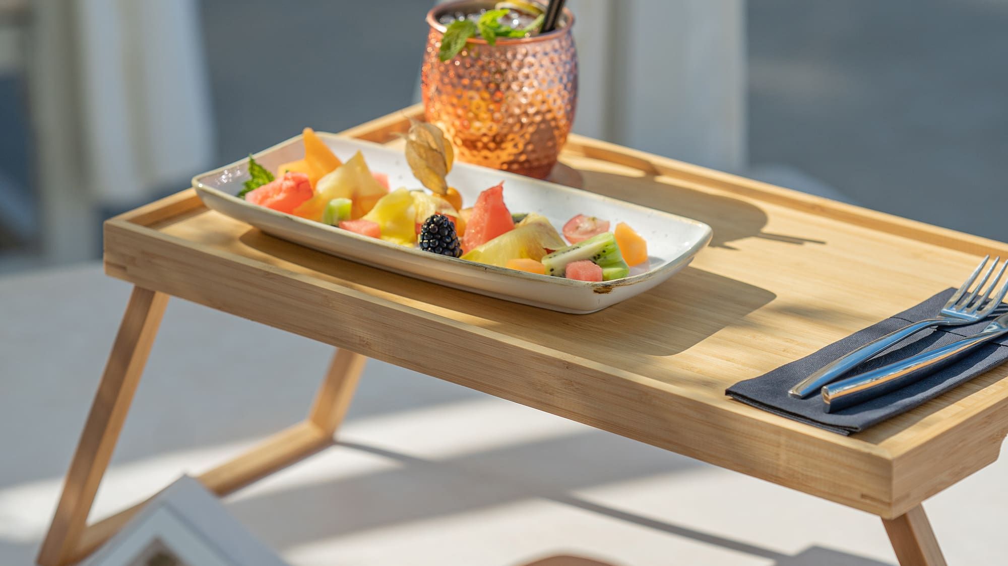 Tropical fruit platter & cocktail on bamboo tray, person reading poolside.