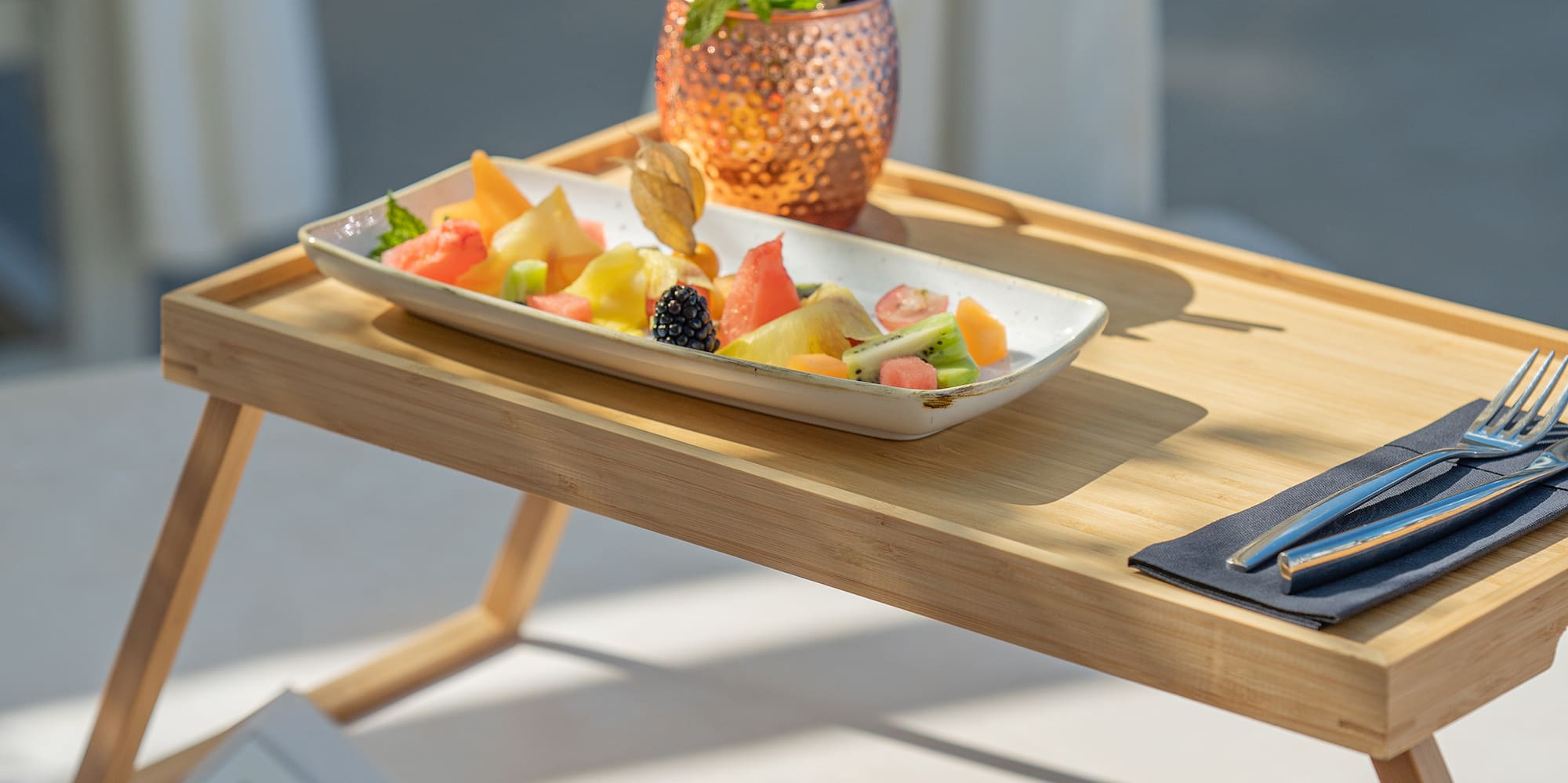 Tropical fruit platter & cocktail on bamboo tray, person reading poolside.