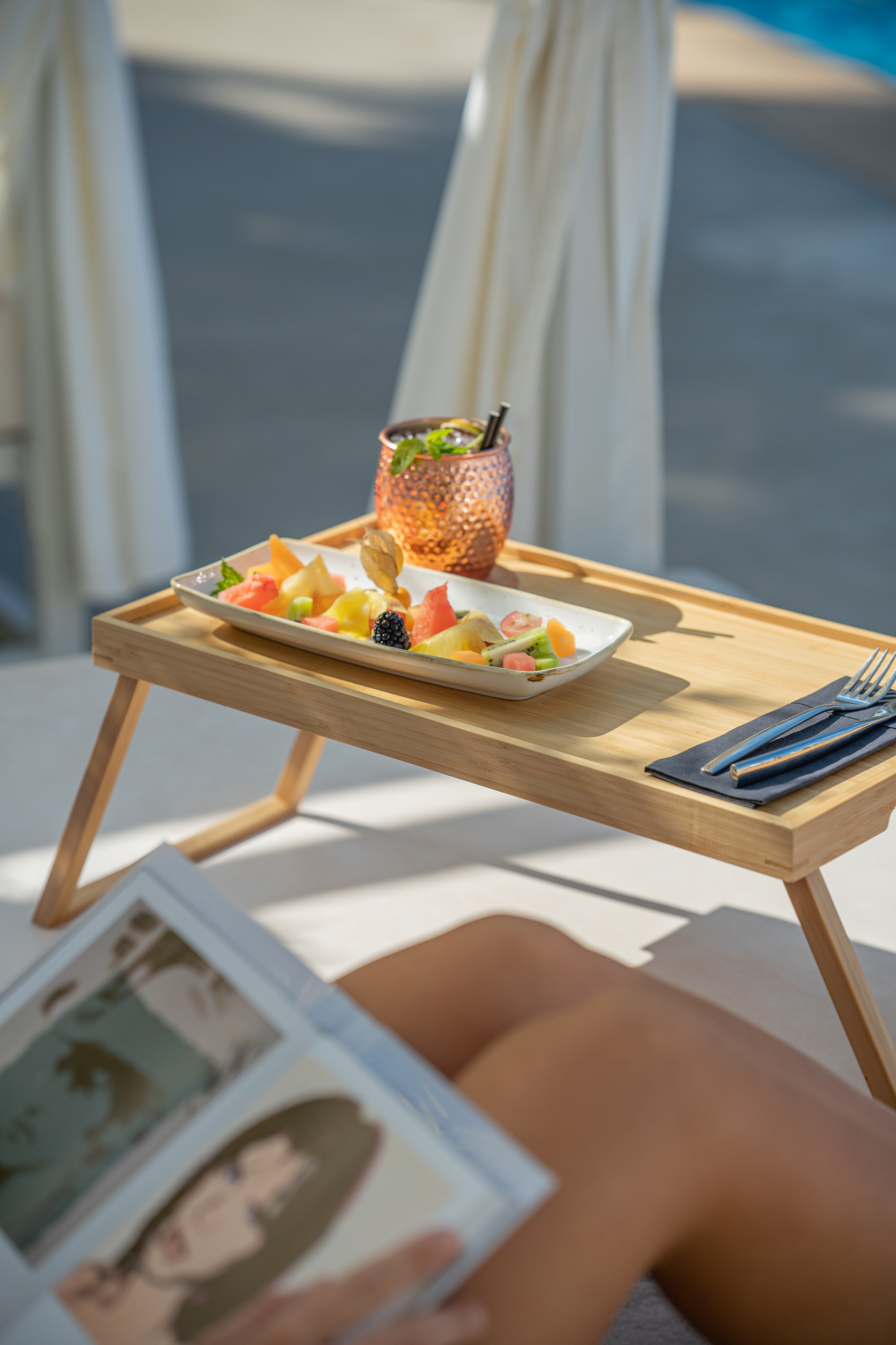 Tropical fruit platter & cocktail on bamboo tray, person reading poolside.