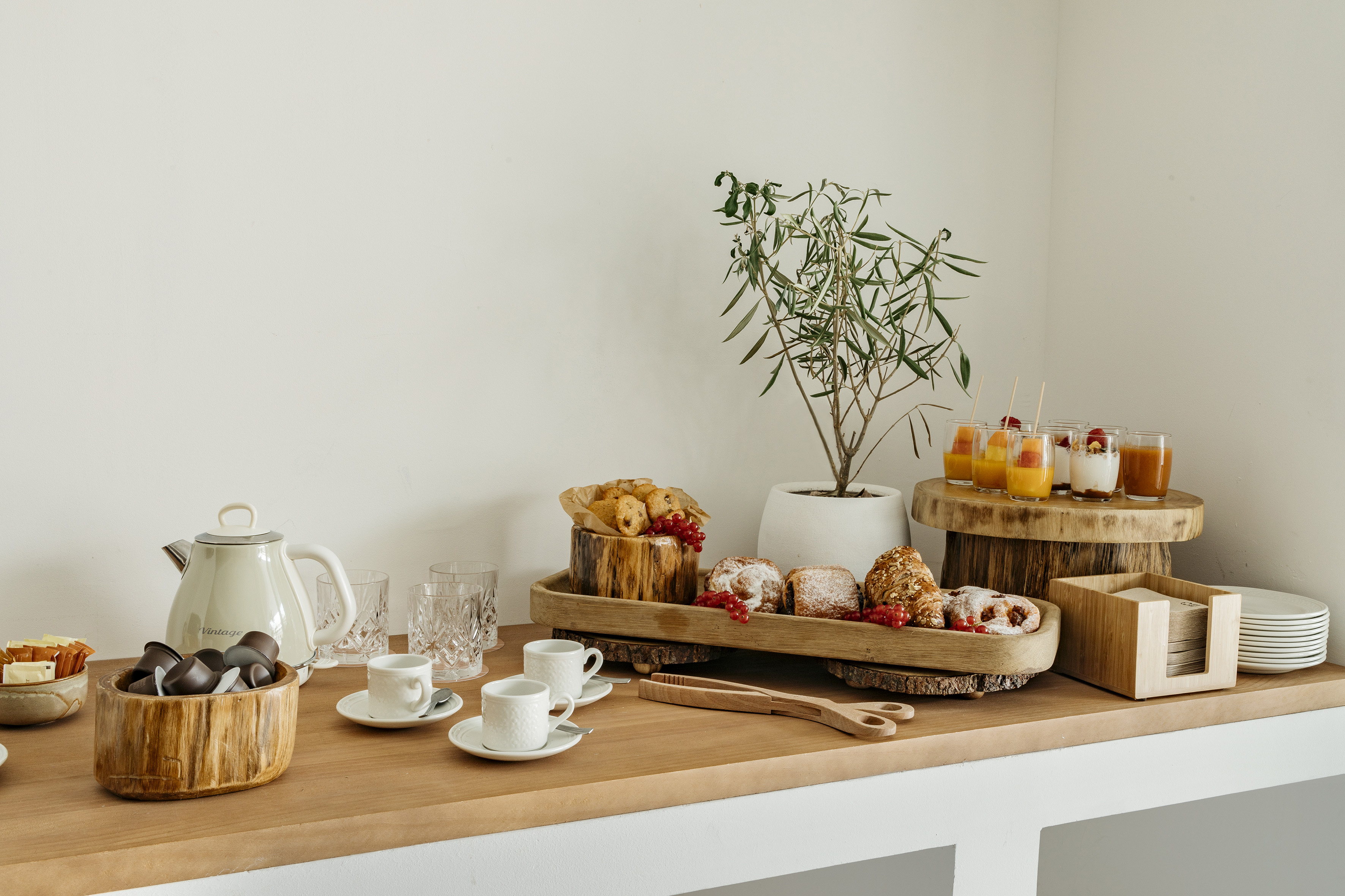 Breakfast spread with pastries, fruit parfaits, coffee, and kettle on wooden counter.