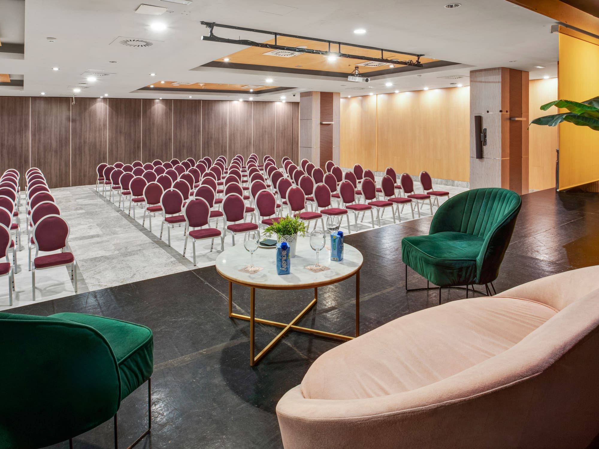 Modern hotel conference room with rows of red chairs, stage, and velvet lounge seating.