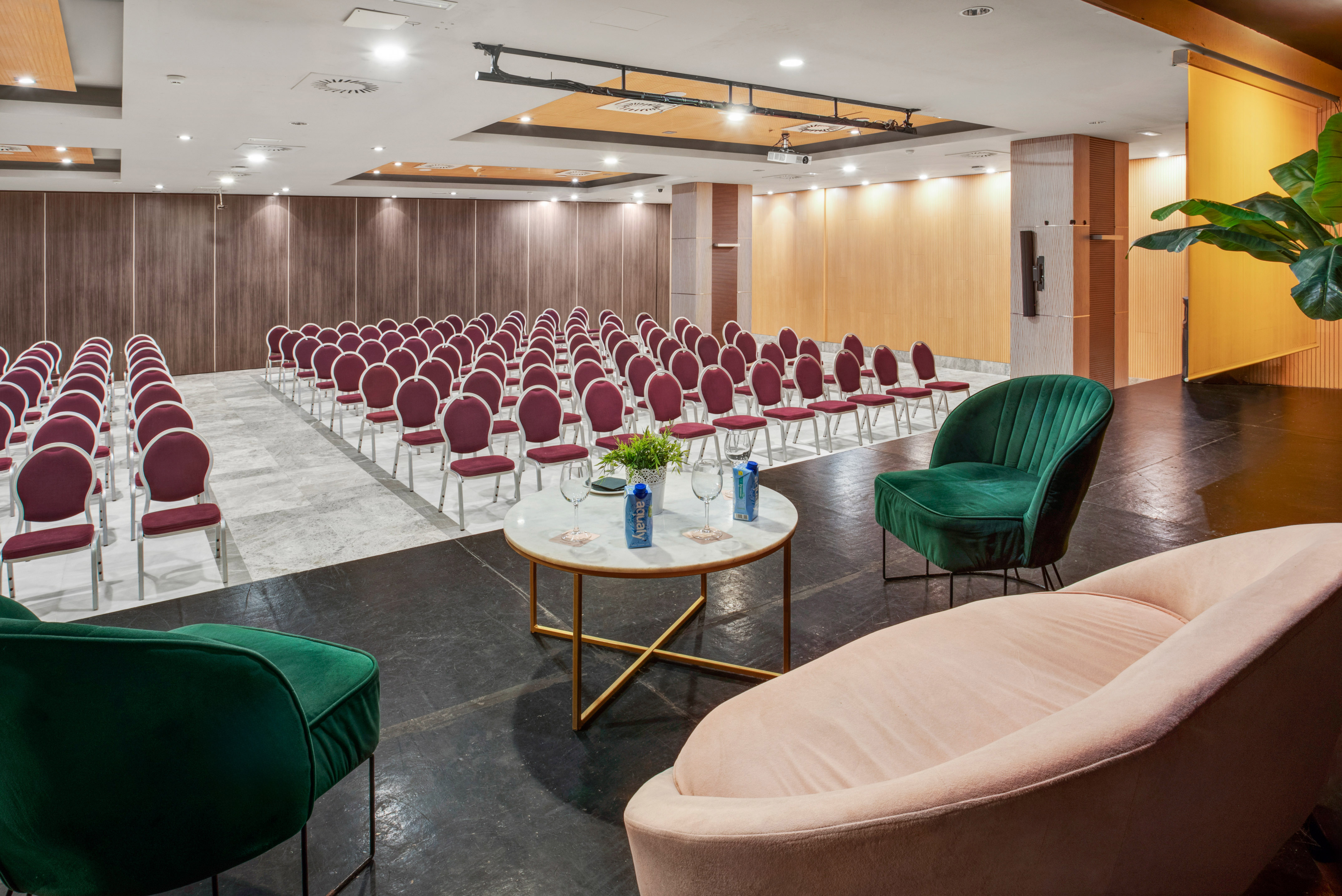 Modern hotel conference room with rows of red chairs, stage, and velvet lounge seating.