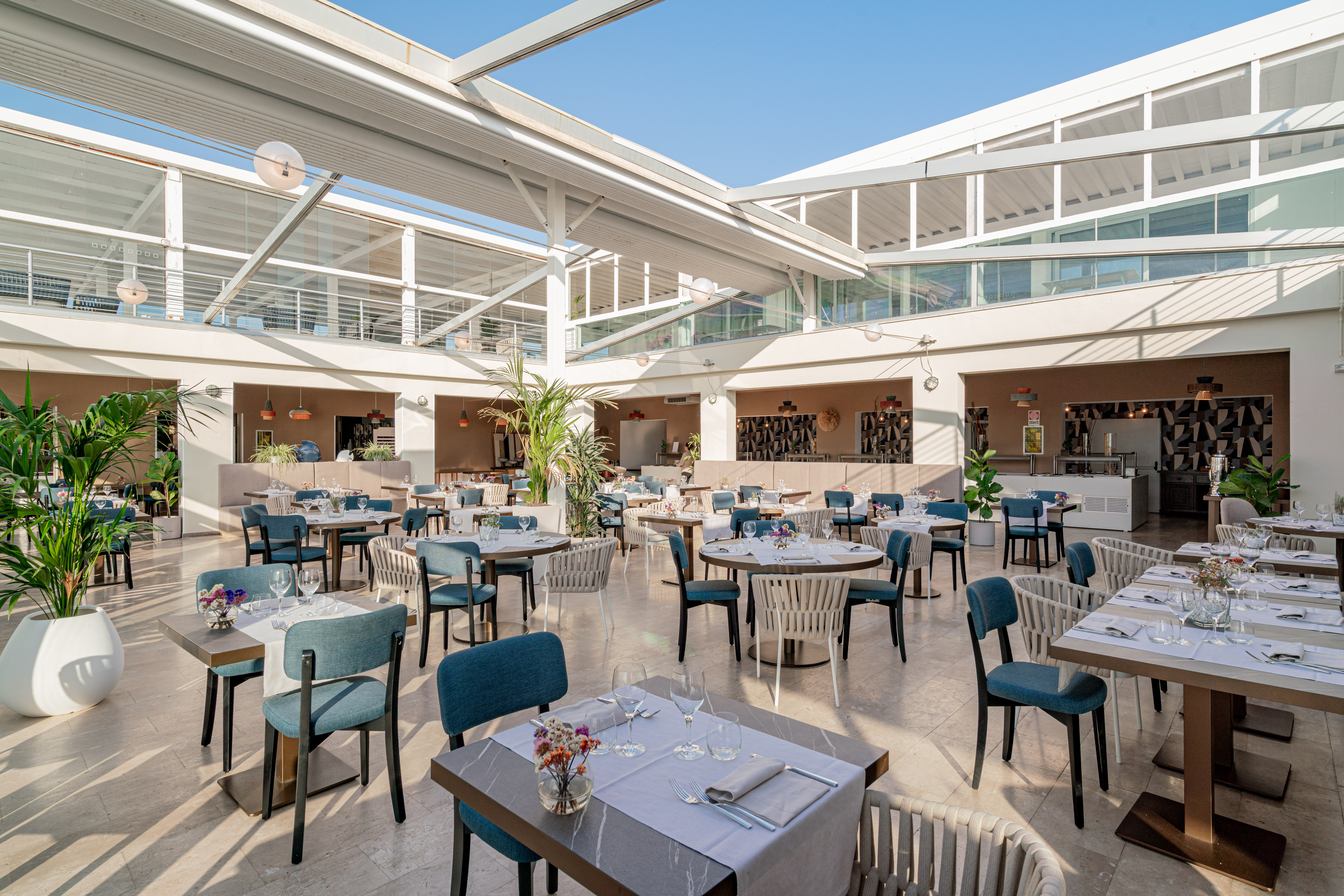 Bright, open-plan hotel restaurant with tables, blue chairs, and skylight.