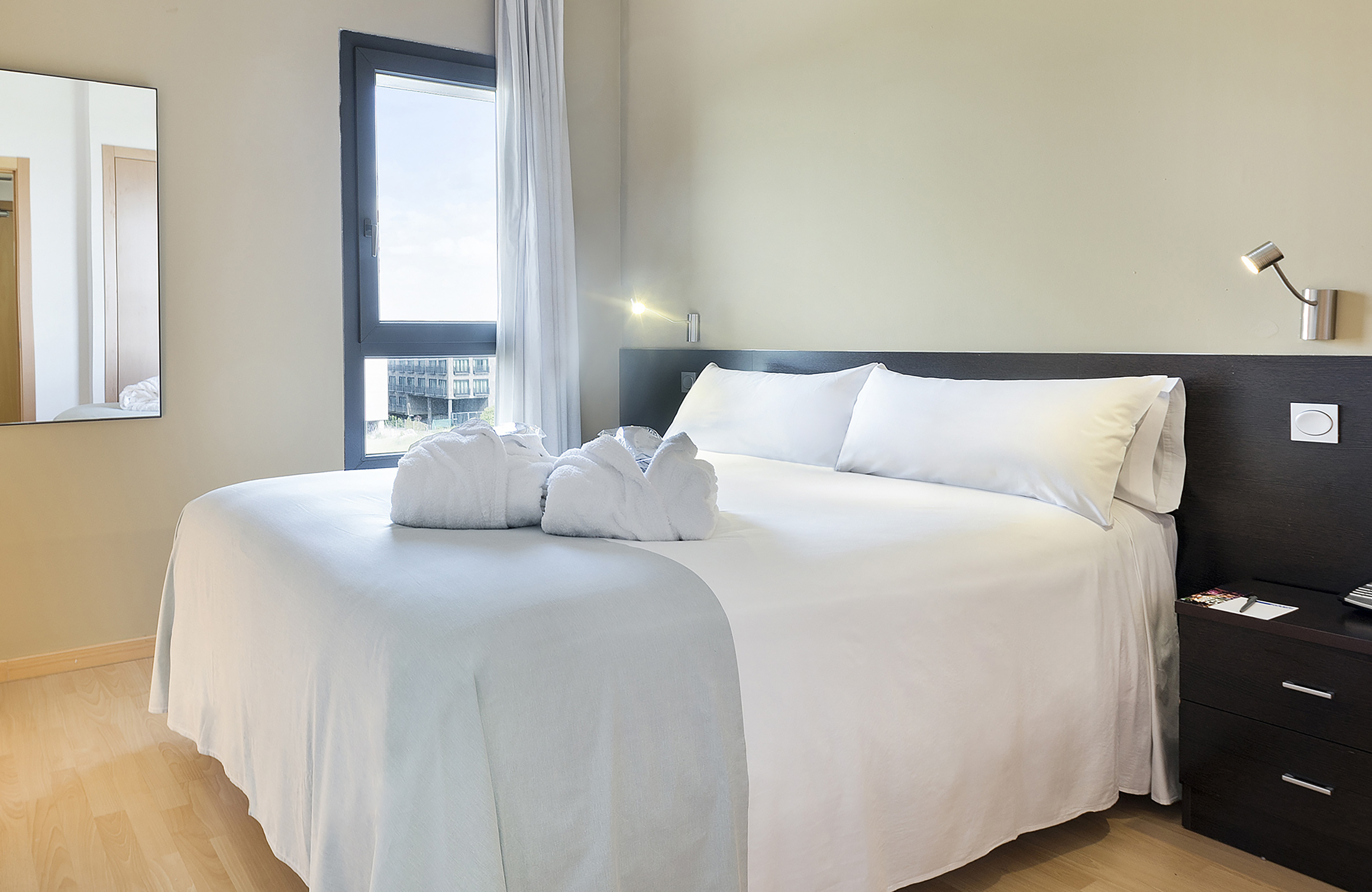 Bright hotel room with queen bed, white bathrobes, city view window.