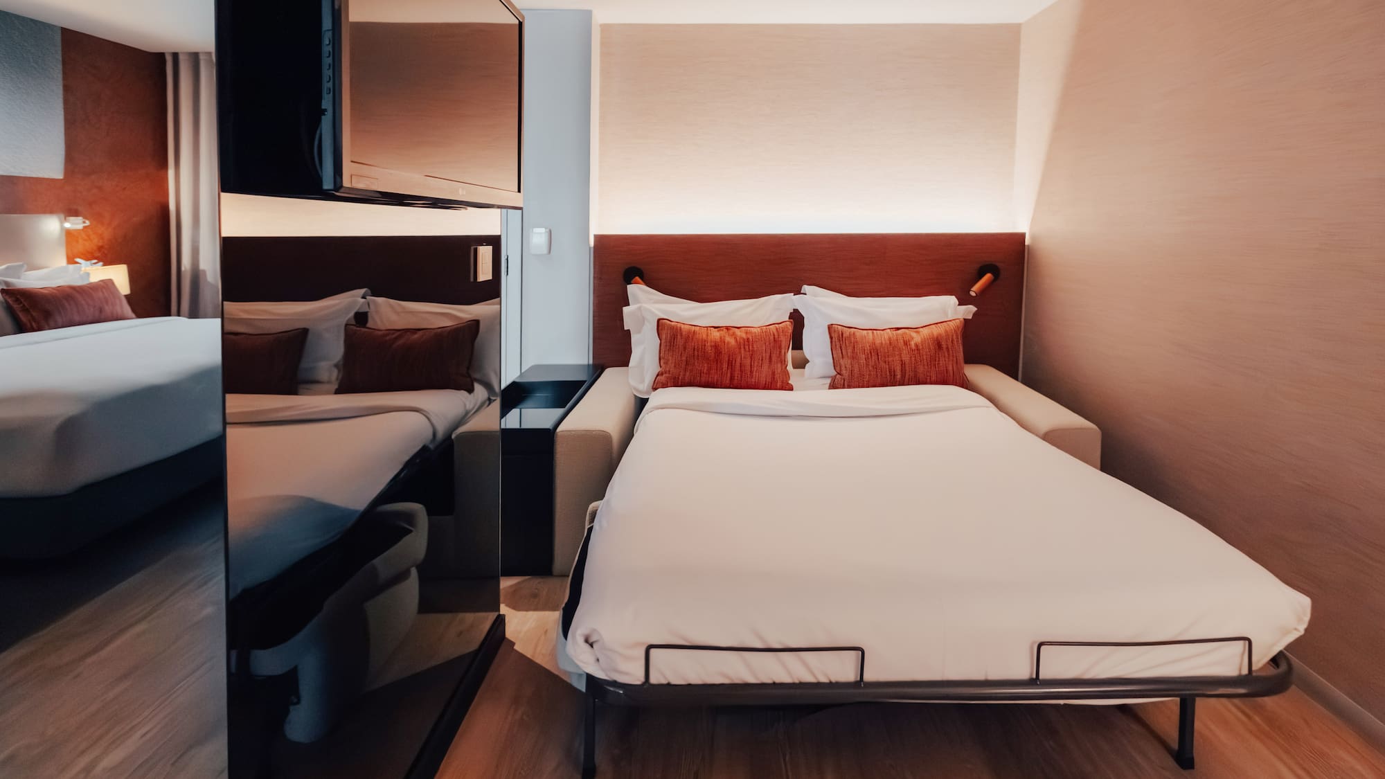 Modern hotel room with pull-out bed, white bedding, and orange pillows.