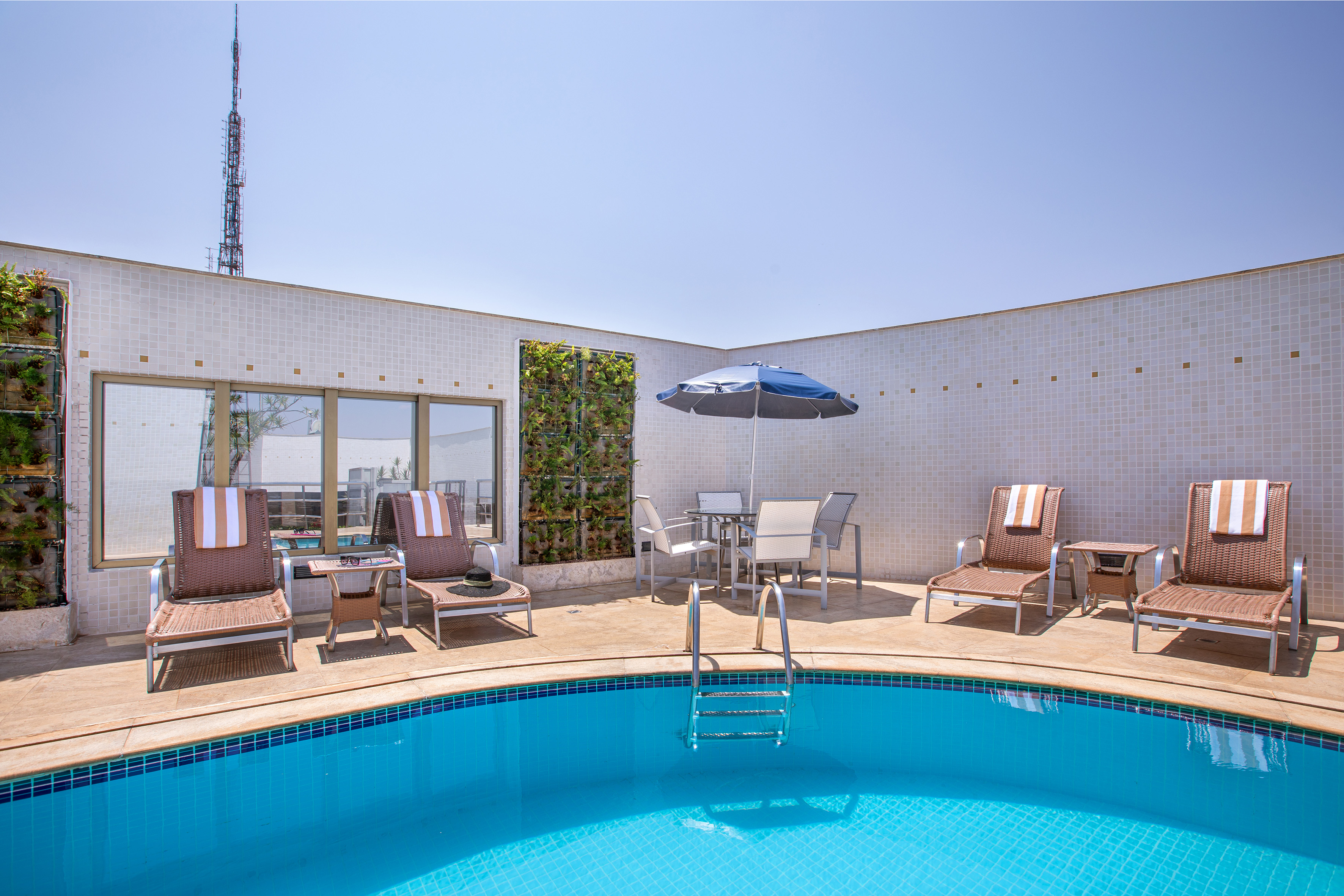 Sunny hotel pool with lounge chairs, patio umbrella, and green wall.