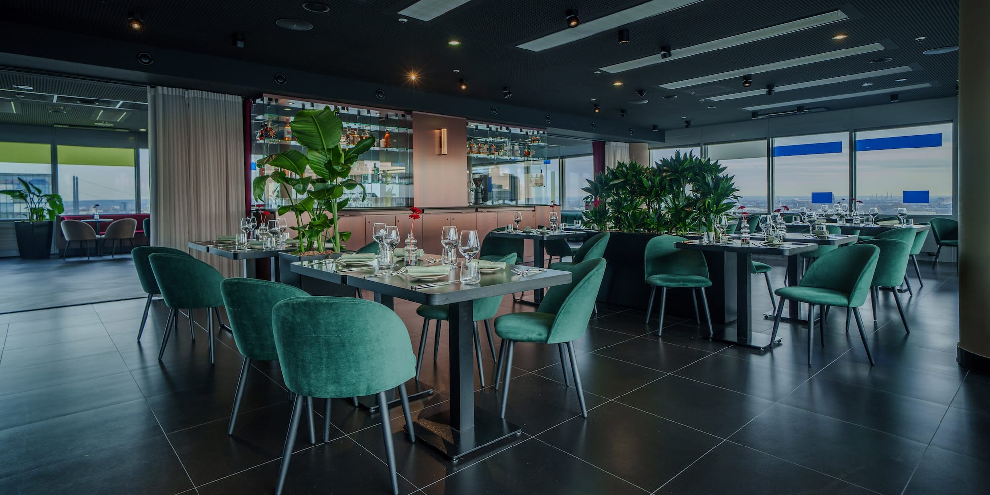 Contemporary restaurant with teal velvet dining chairs, bar, and city view.