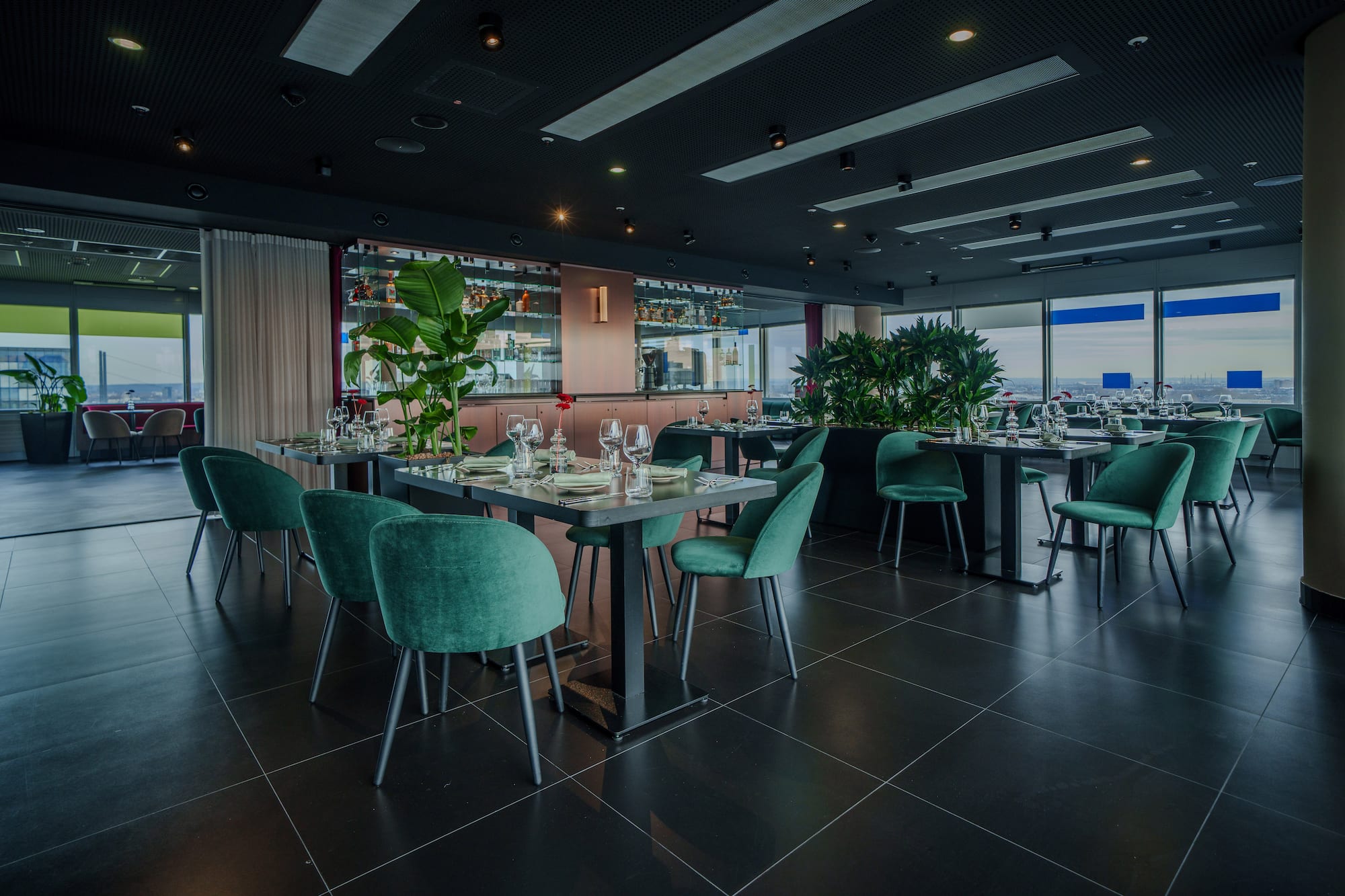 Modern restaurant interior with green velvet chairs, bar, and cityscape views.