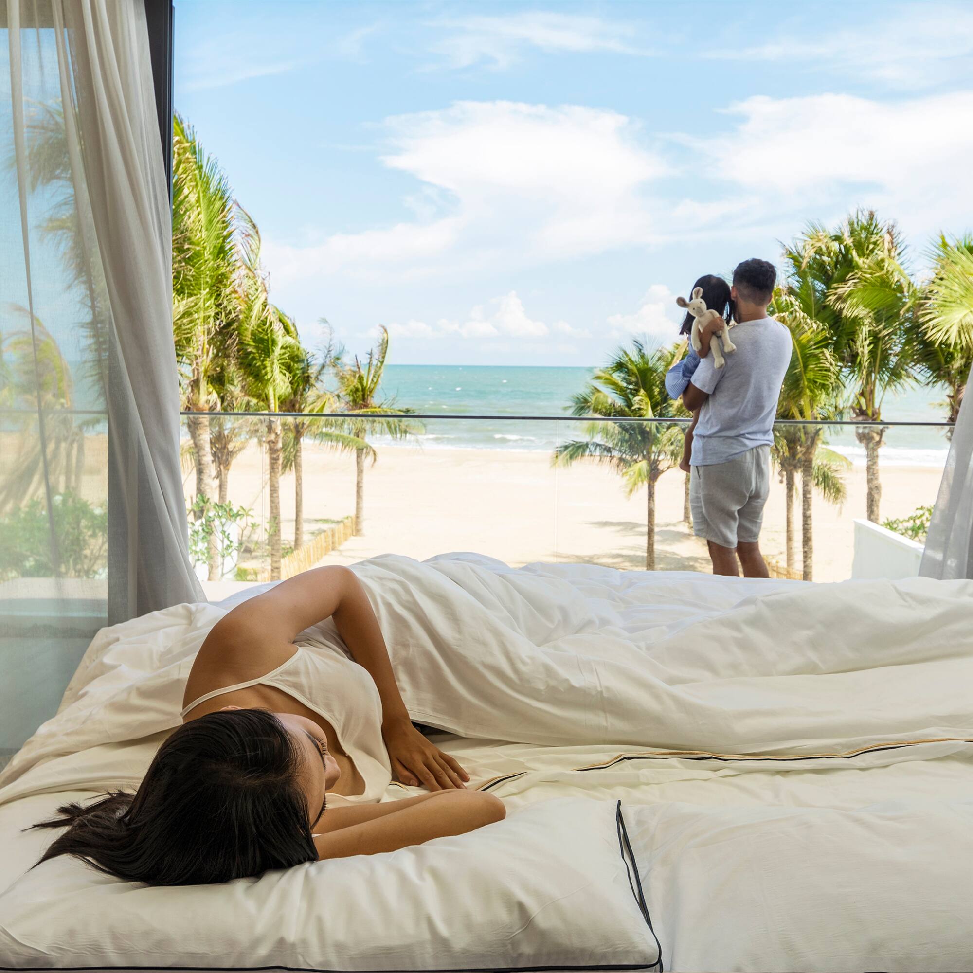 Woman in bed, family on balcony overlooking beach and palm trees.