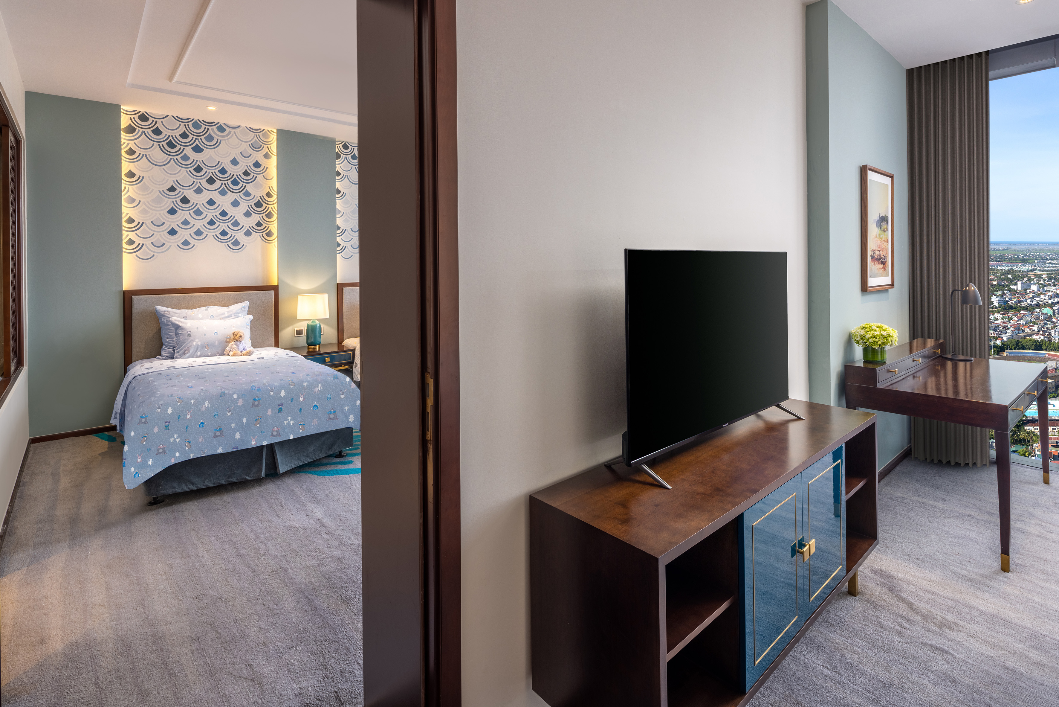 Modern hotel suite with kids' bedroom, TV, desk, and city view.
