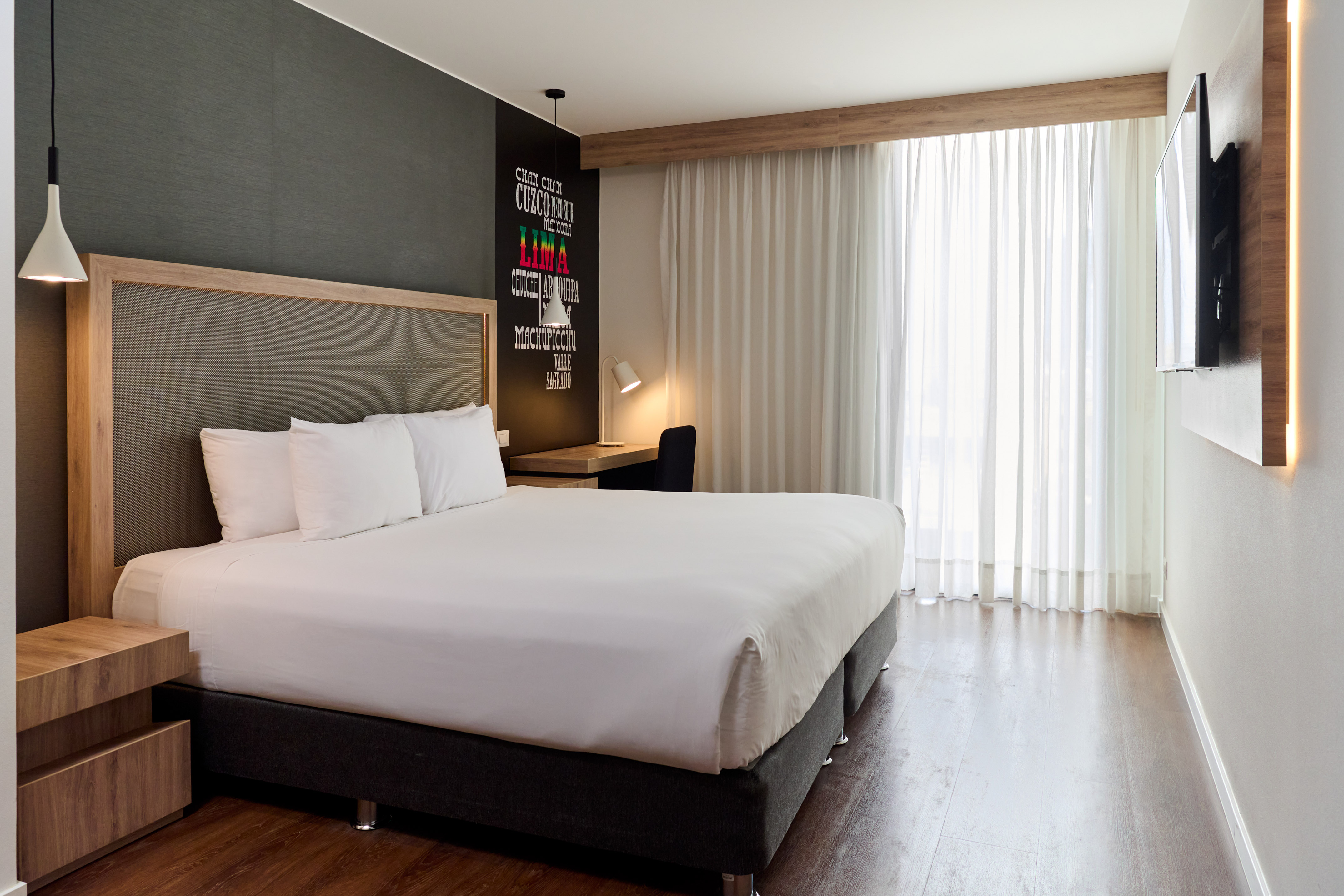 Modern hotel room with bed, desk, city names wall art, and bright window.
