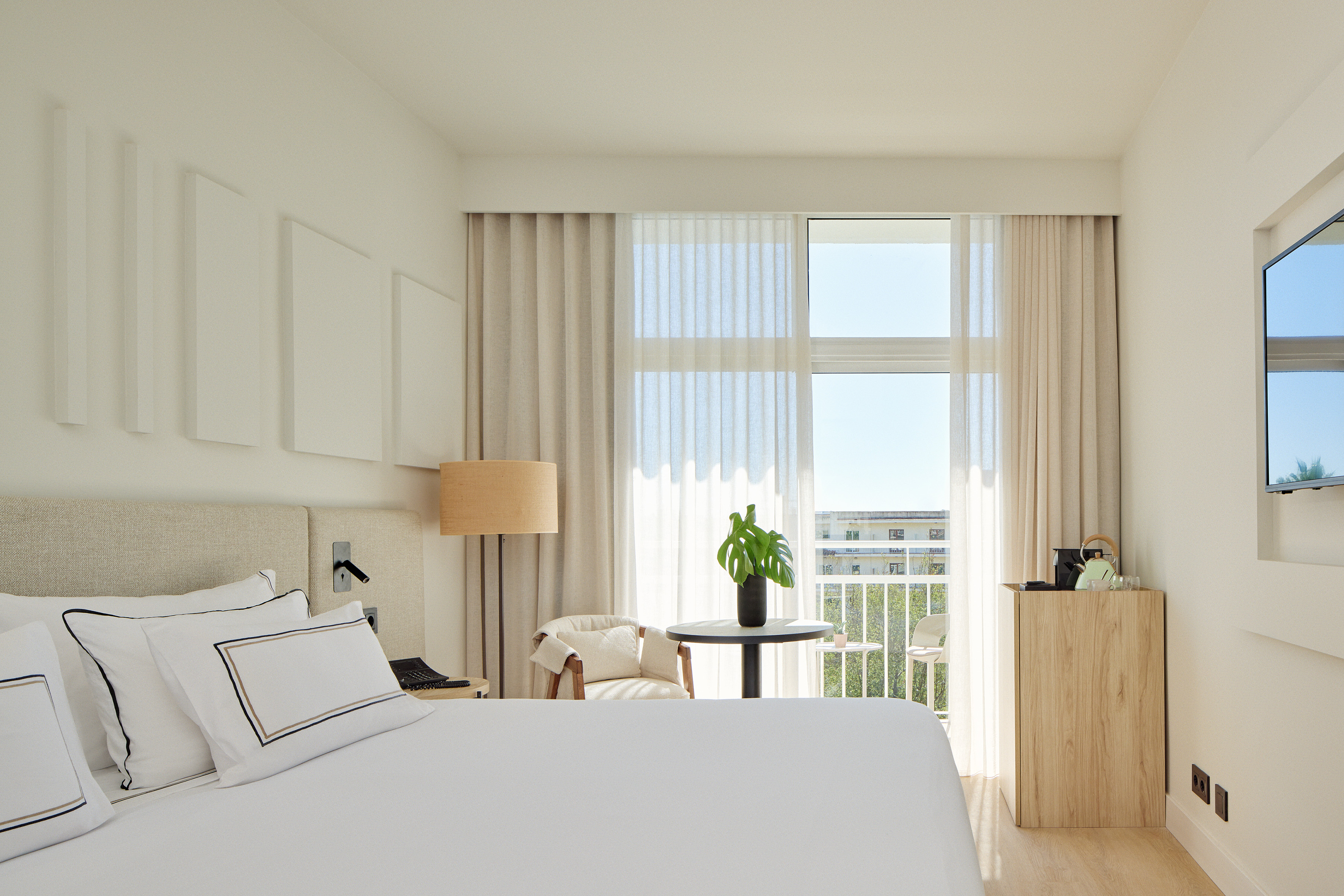 Modern hotel bedroom with white bed, bright balcony, and natural light.