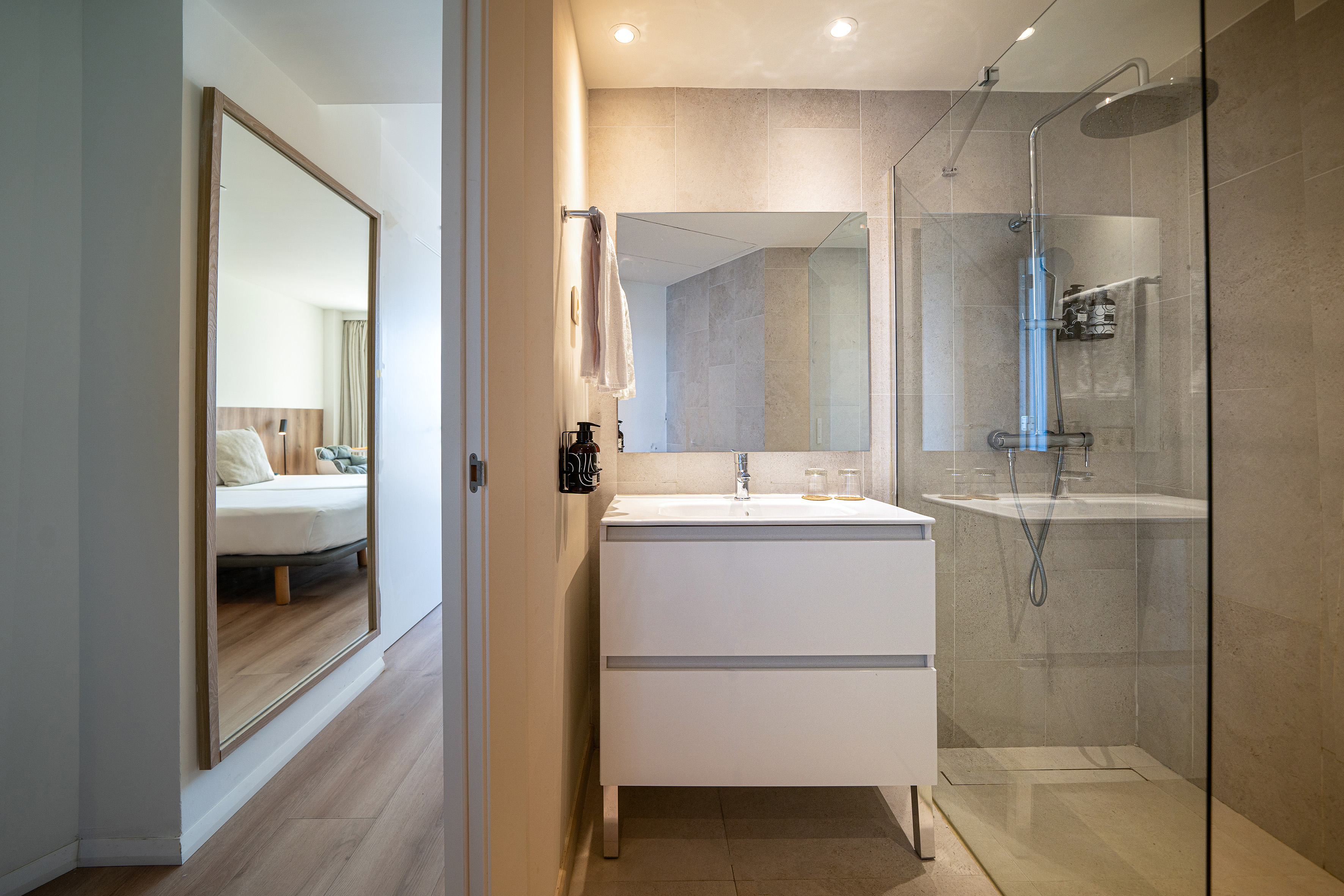 Modern hotel bathroom with walk-in shower, vanity, mirror reflecting bedroom.
