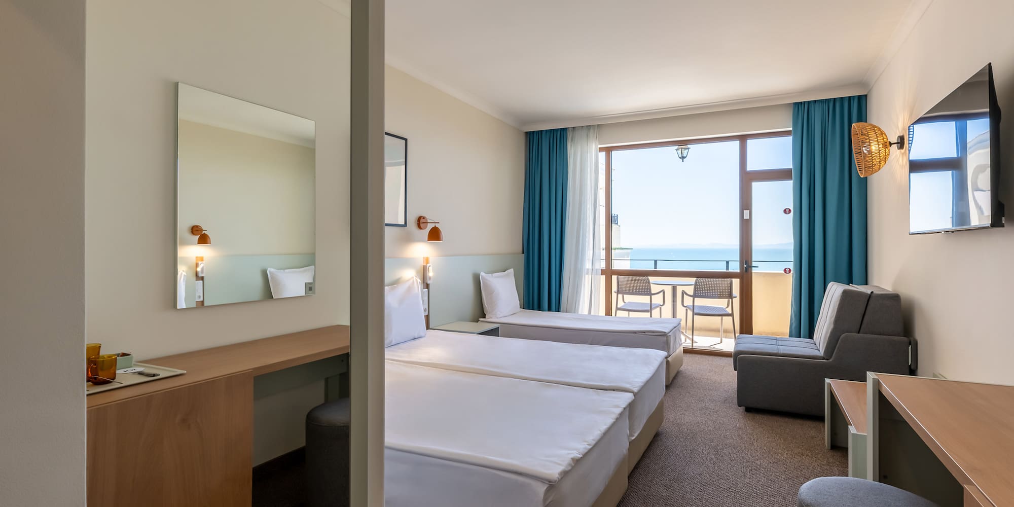 Modern hotel room with twin beds, ocean view balcony, desk, and sofa.